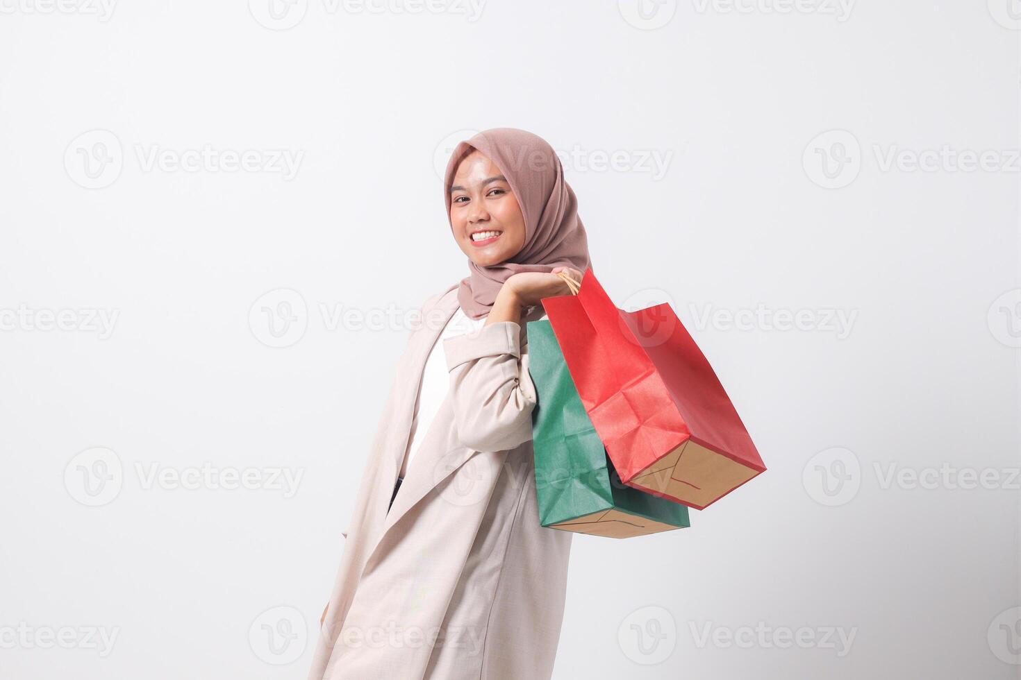 Portrait of cheerful Asian hijab woman in casual suit holding and showing colorful paper shopping bags. Shopaholic girl and discount buying concept. Isolated image on white background photo