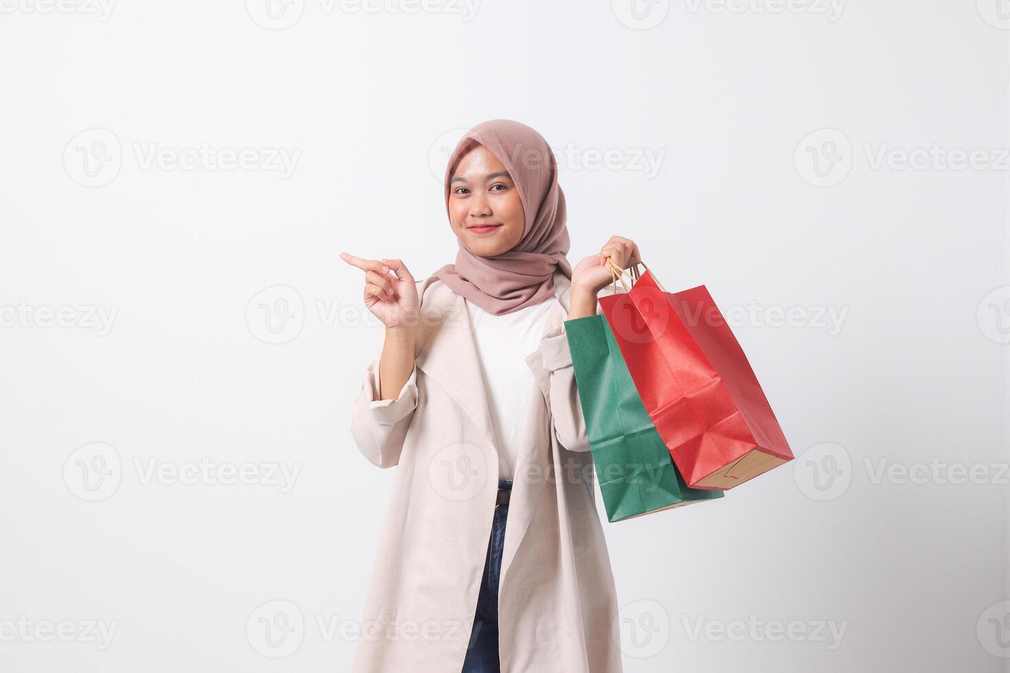 Portrait of excited Asian hijab woman in casual suit holding paper shopping bags while pointing with finger. Shopaholic girl and discount buying concept. Isolated image on white background photo