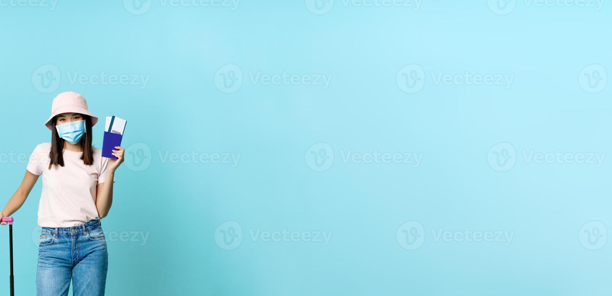 Vertical shot of happy asian woman with suitcase, showing passport and two tickets, wearing face mask from coronavirus, blue background photo