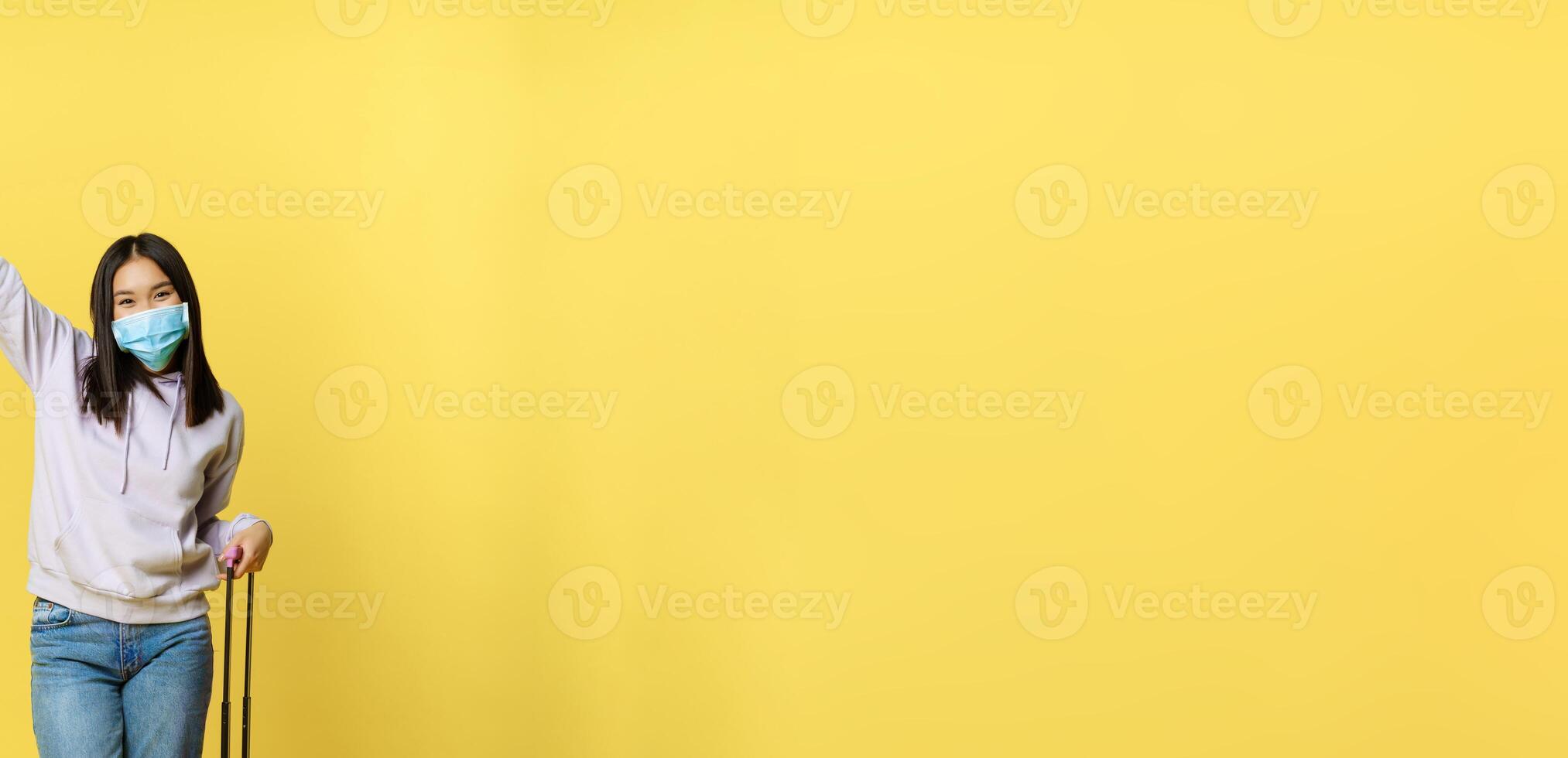Full length shot happy asian woman going on vacation, tourist with suitcase posing cheerful, wearing medical face mask, yellow background photo