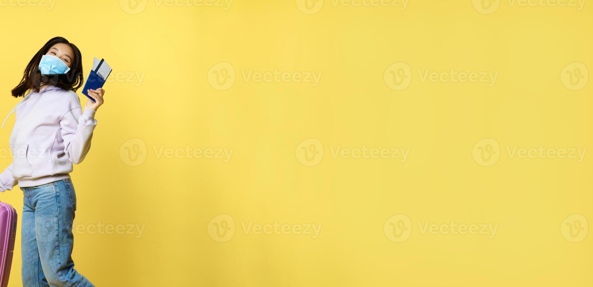 Full length shot happy asian woman jumping with suitcase and tickets on tour, holding passport, going on vacation, standing over yellow background photo