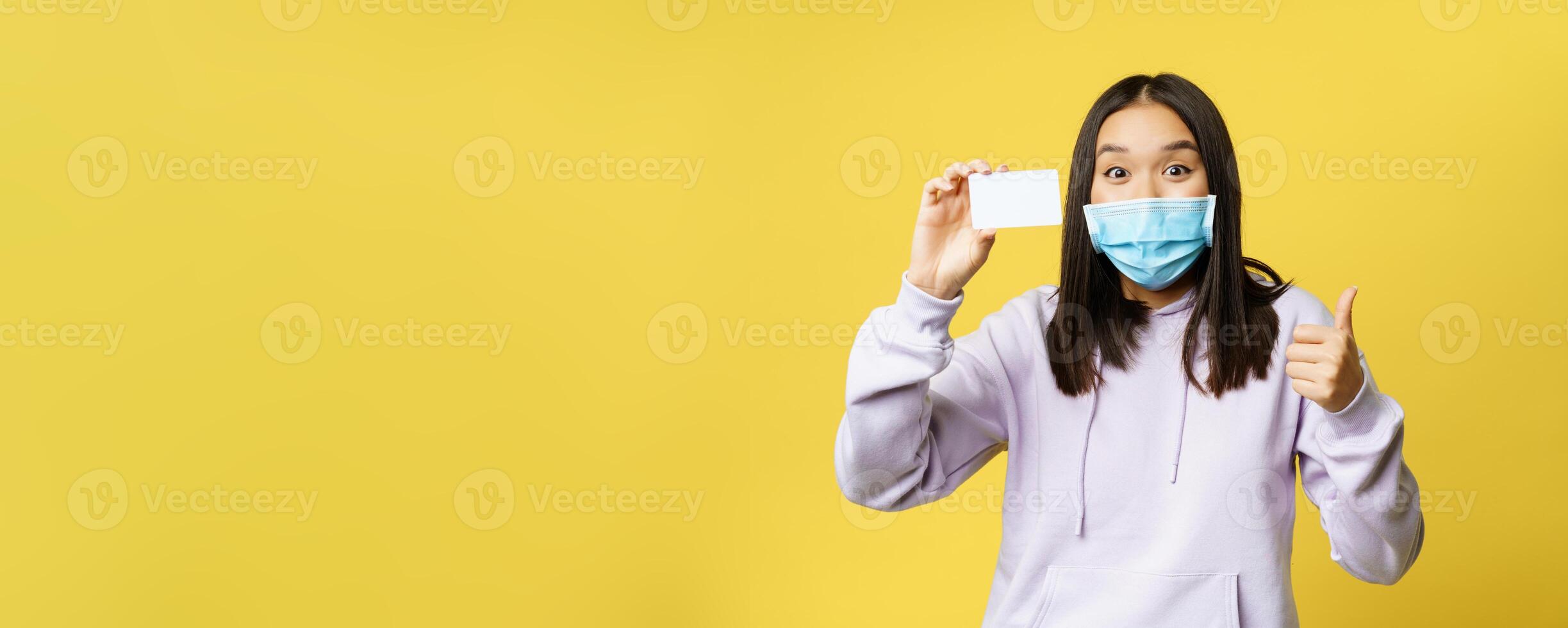 Enthusiastic korean girl in face mask, showing card and thumbs up, recommending product, shopping, standing over yellow background photo