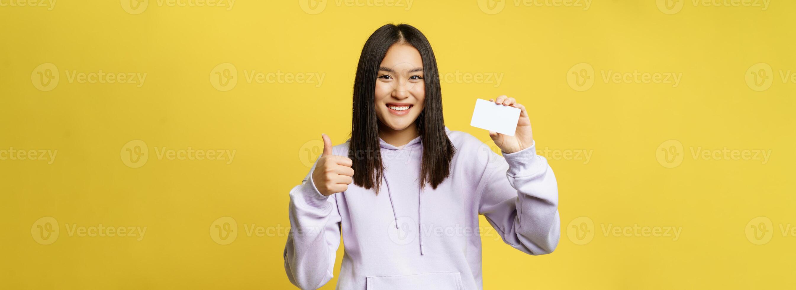 Smiling japanese girl showing card and looking pleased, thumbs up gesture, standing over yellow background photo