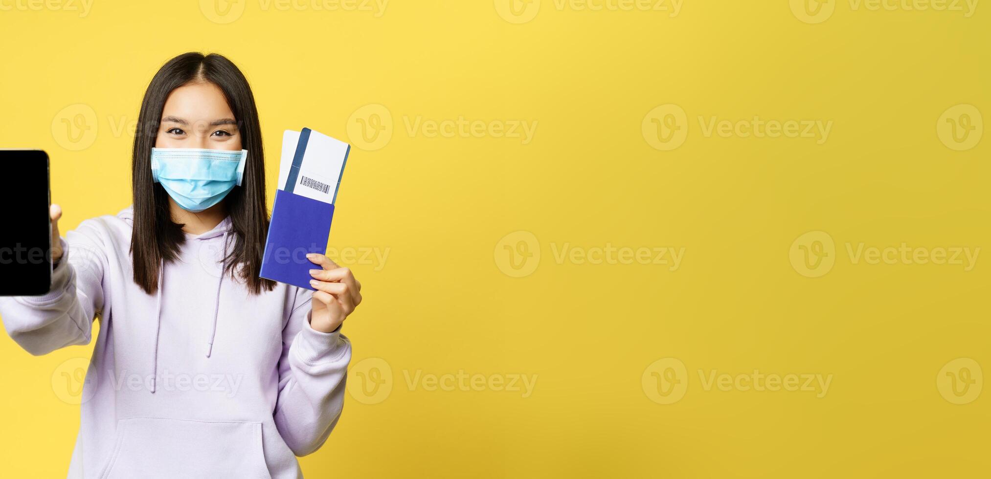 Happy korean girl in face mask, showing smartphone screen, app interface and passport with tickets for vacation, booking trip, yellow background photo