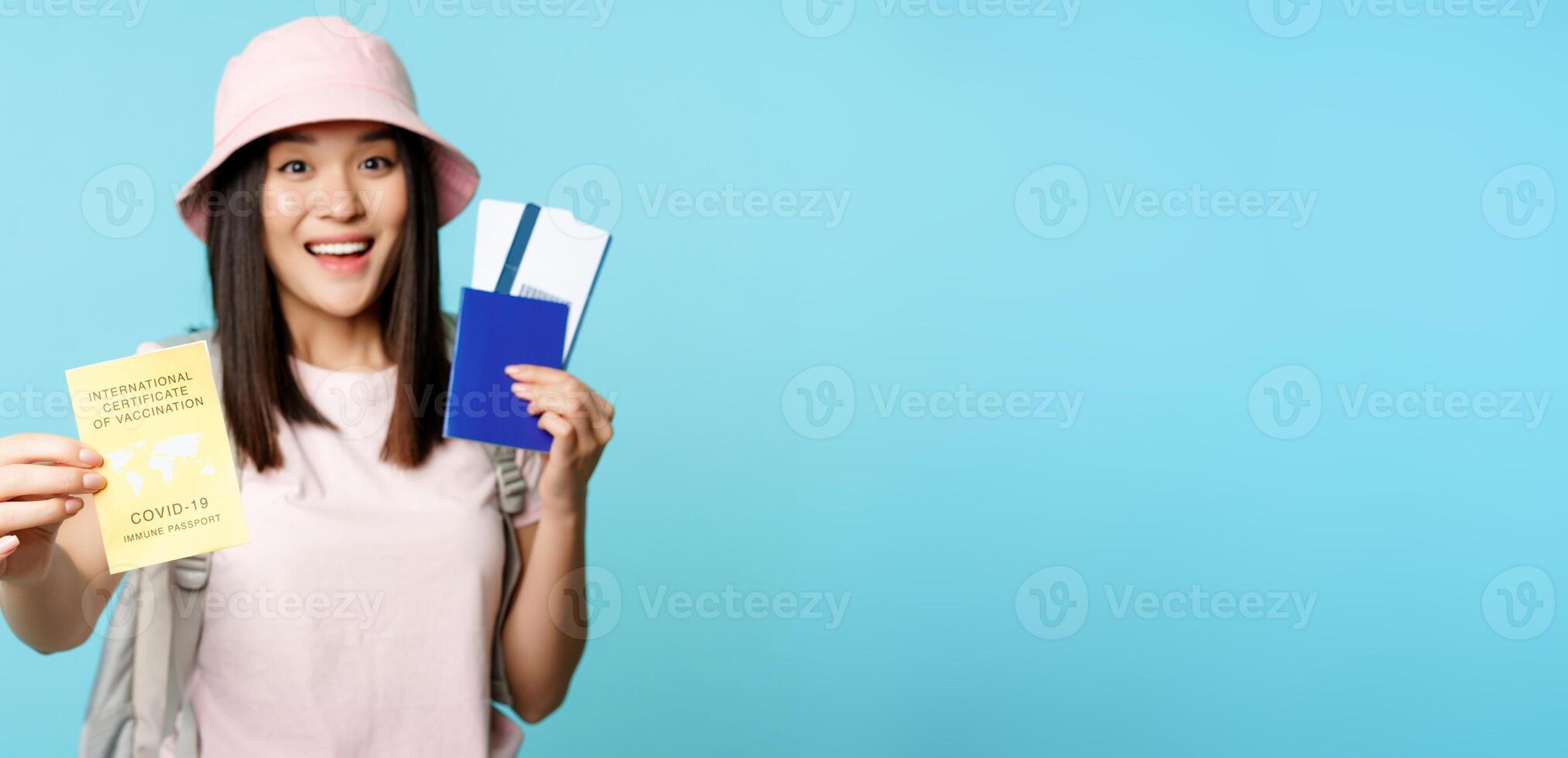 Enthusiastic asian tourist, girl shows covid-19 international vaccination certificate, passport with two tickets, travelling after coronavirus vaccine, blue background photo