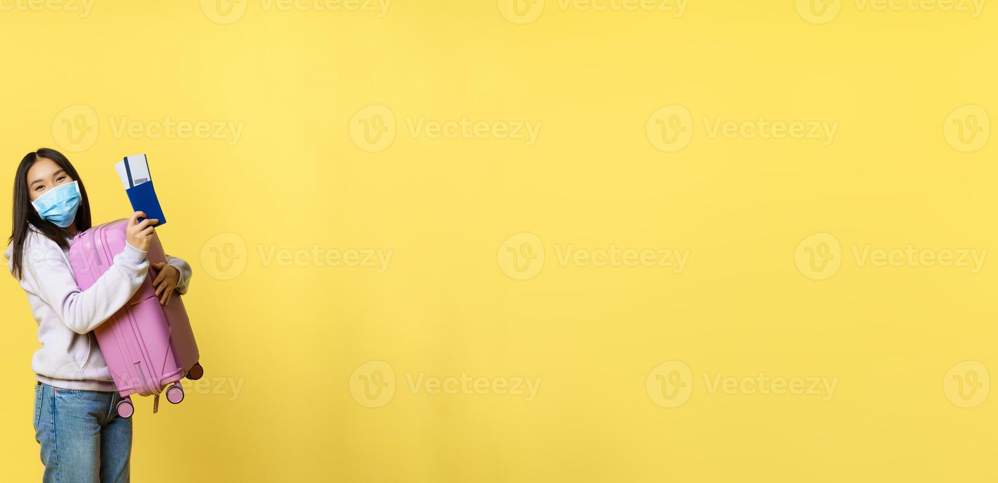 Full length shot cute korean girl tourist in medical face mask, holding her suitcase and passport with flight tickets, going on vacation abroad, yellow background photo