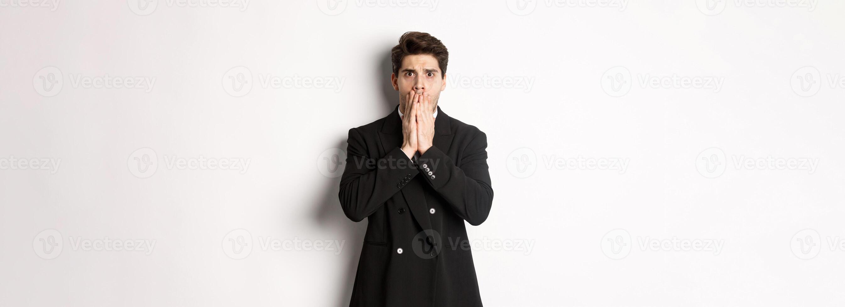 Portrait of shocked handsome businessman in suit, reacting to terrible situation, gasping and covering mouth with hands, standing startled against white background photo