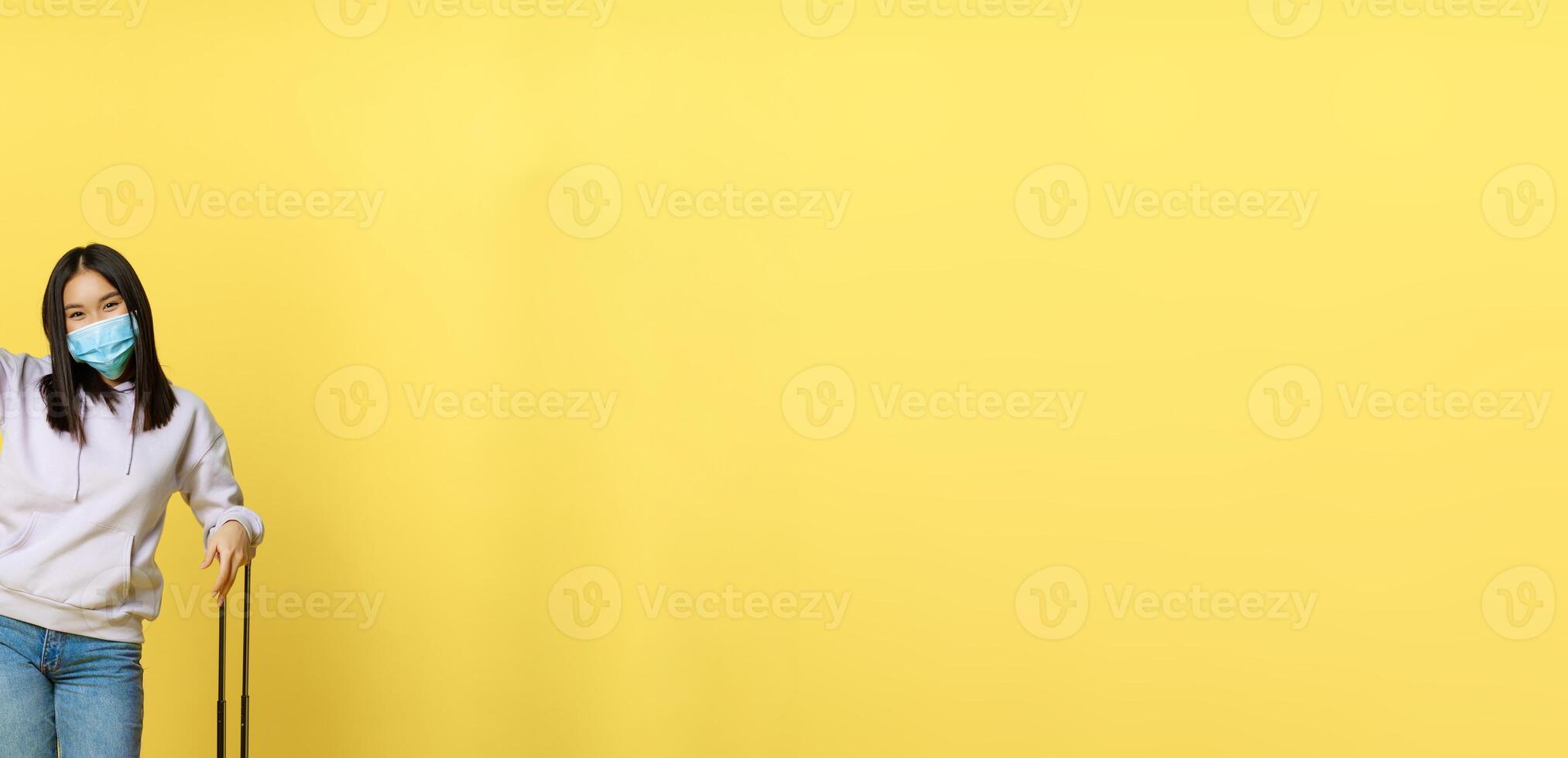 Full length shot of enthusiastic korean girl enjoying vacation, posing with suitcase, wearing face medical mask, travelling abroad during covid pandemic, yellow background photo