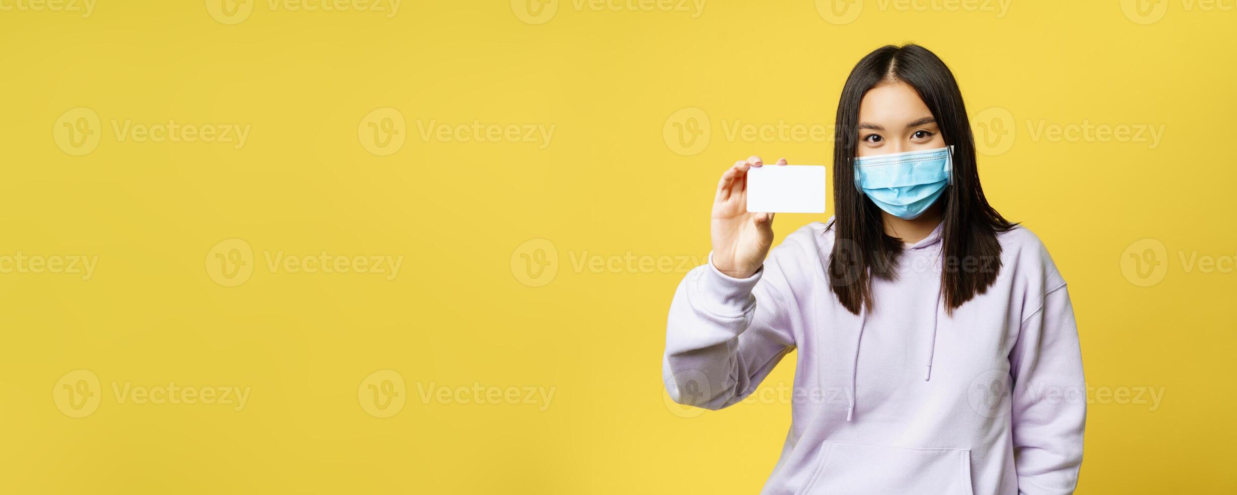 Smiling korean girl in face mask, showing card, standing in casual hoodie over yellow background photo