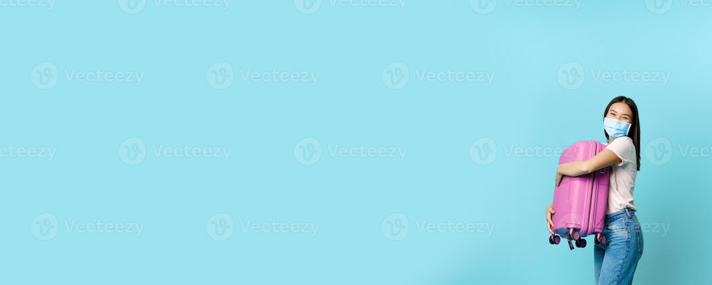 Full length of happy asian female tourist, dancing with suitcase in medical face mask, going on vacation, travelling with briefcase, standing over blue background photo