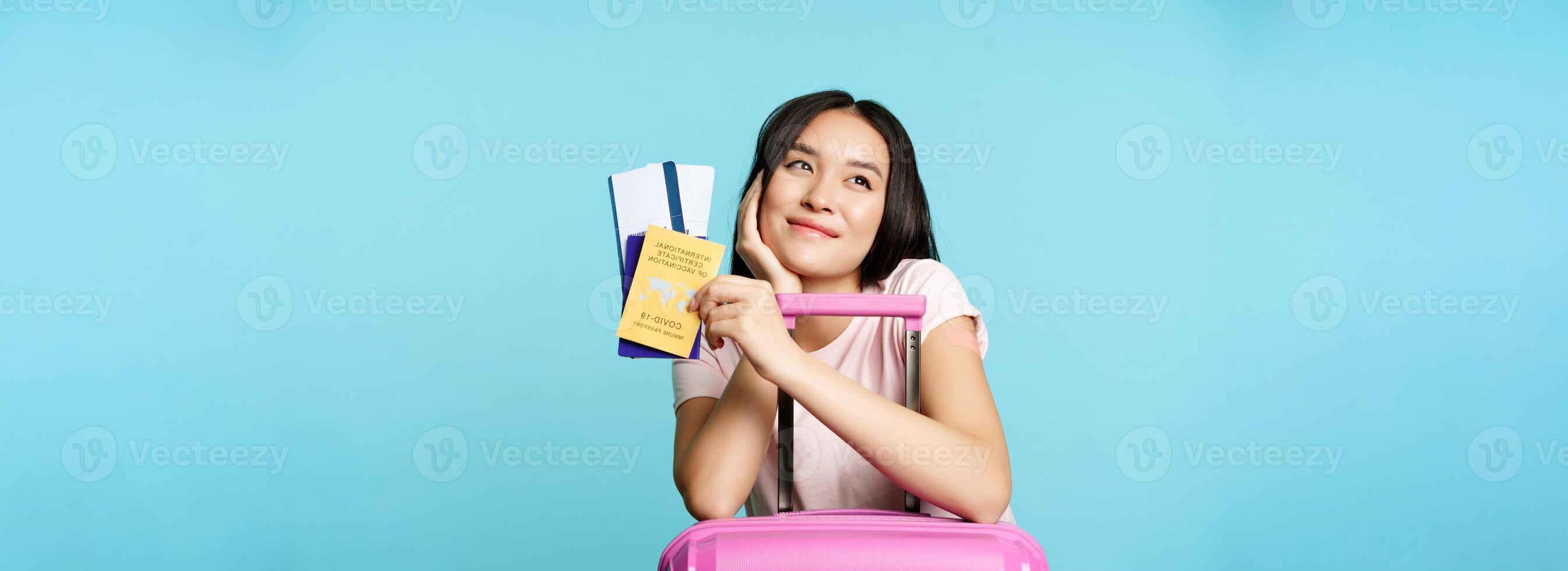 Dreamy asian tourist, smiling and thinking, holding covid-19 international vaccination certificate, two tickets and passport, standing with suitcare for trip, blue background photo