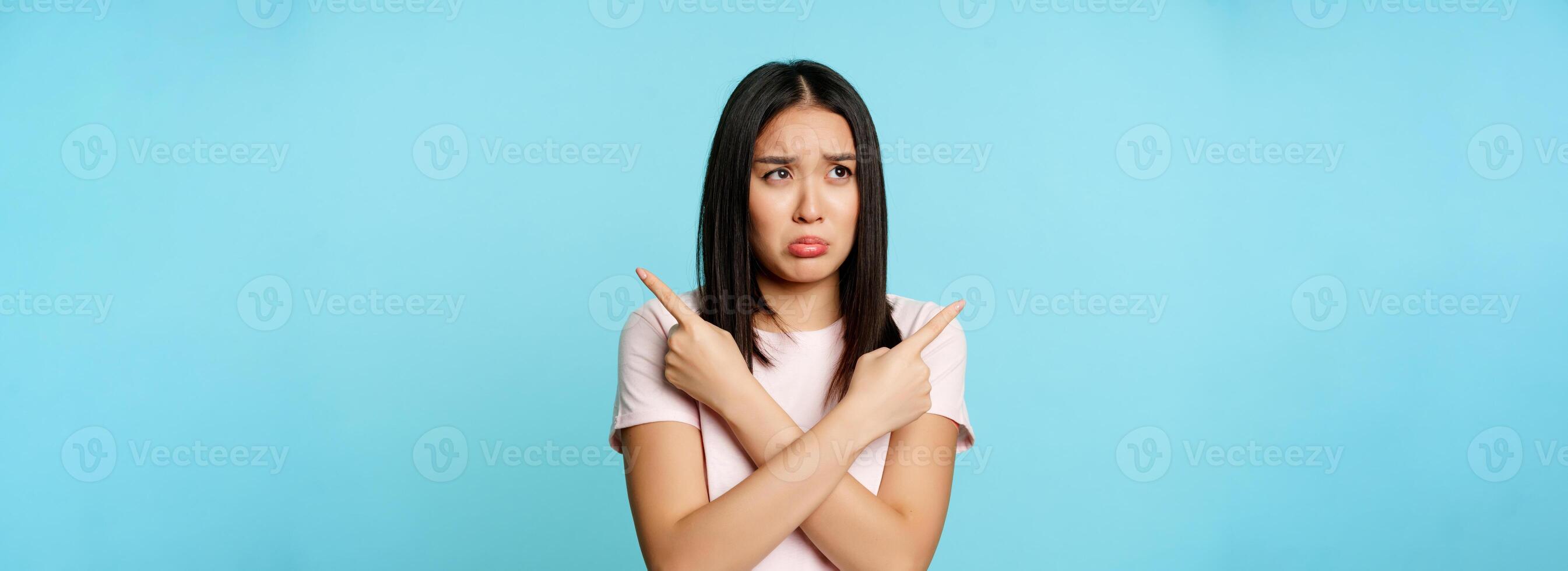 Confused and sad asian girl pointing sideways, looking clueless or indecisive, problem with choosing, pulling long upset face, blue background photo