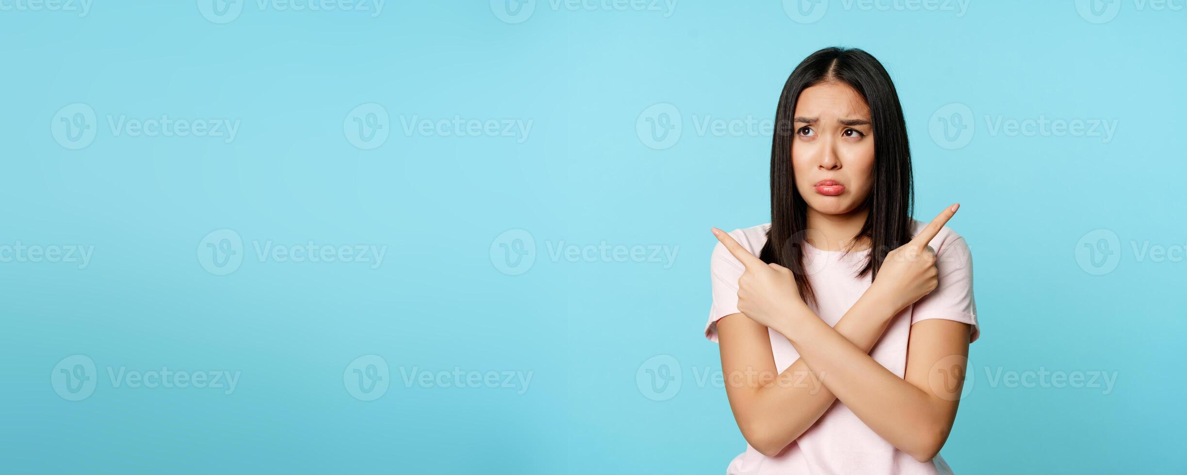 Confused and sad asian girl pointing sideways, looking clueless or indecisive, problem with choosing, pulling long upset face, blue background photo
