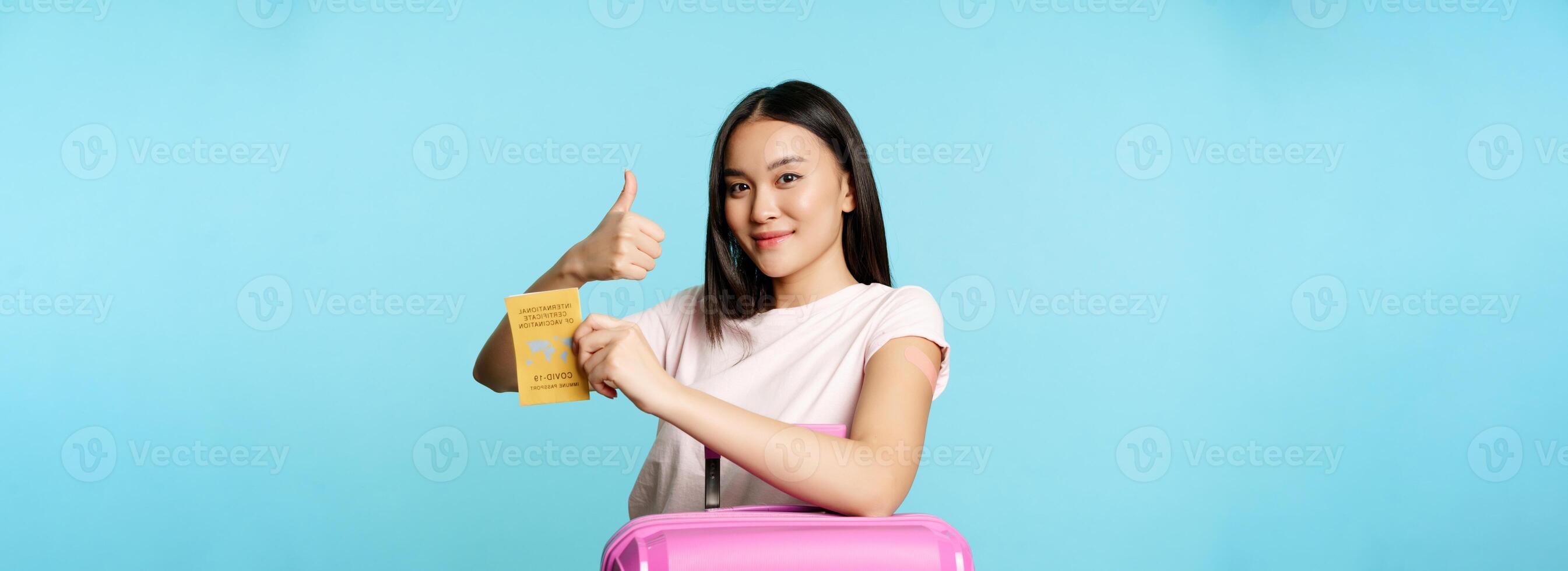 Healthcare and tourism. Smiling asian girl got vaccinated from covid-19, standing with suitcase, showing coronavirus international vaccination passport, thumbs up and arm with patch photo