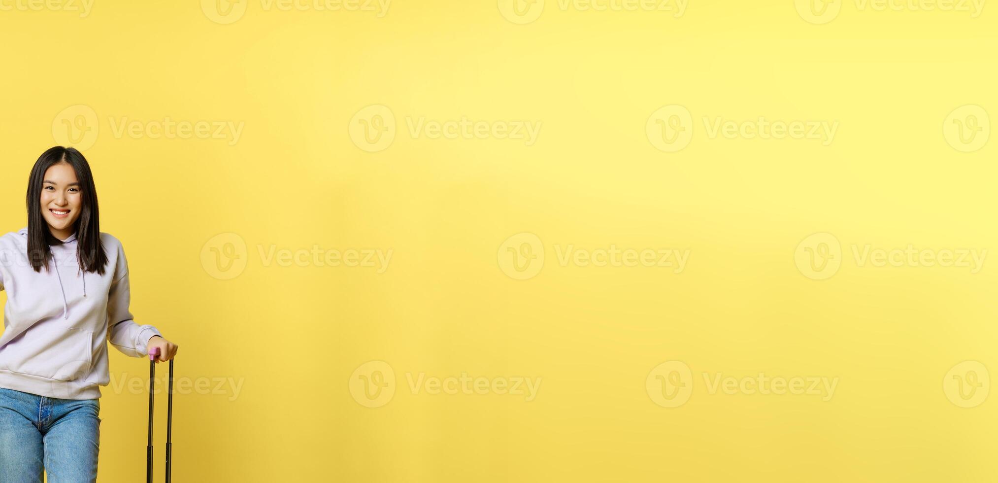 Full length shot of smiling asian tourist, girl traveller pointing at promo, advertisement, standing with suitcase going on vacation, yellow background photo