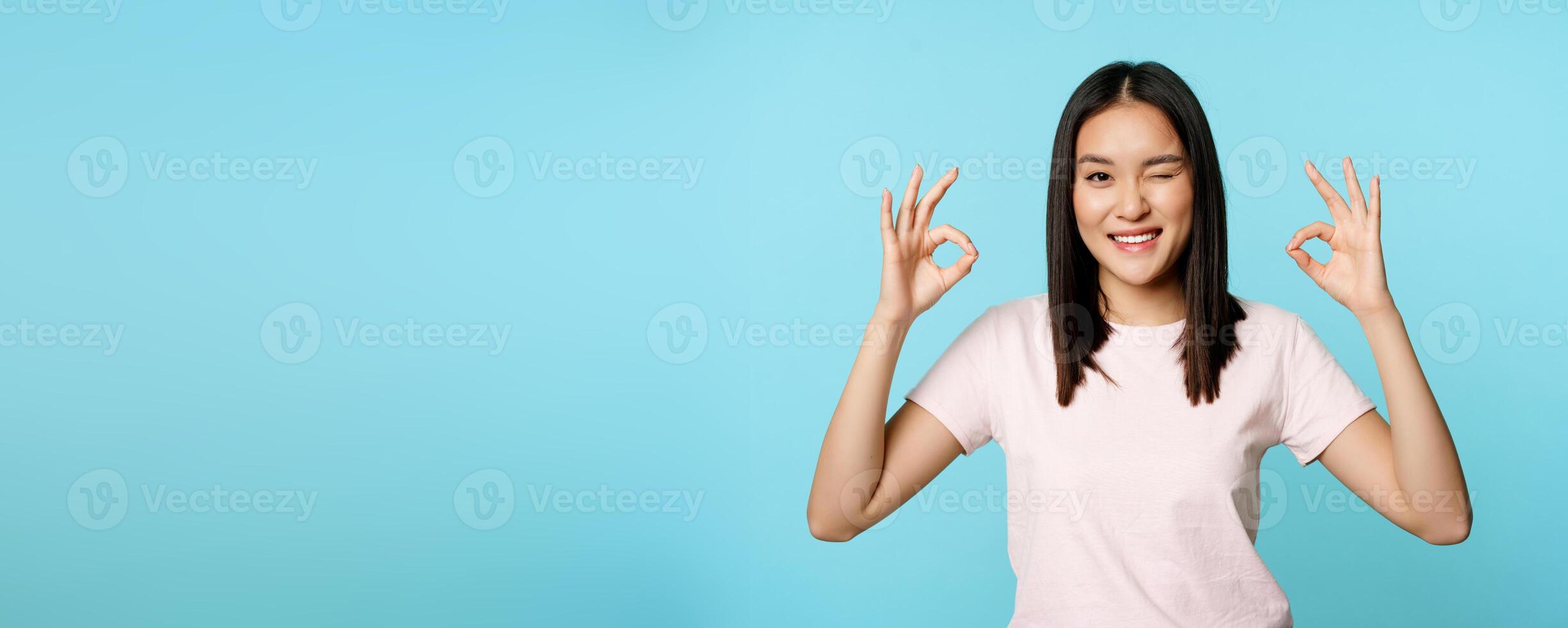 Smiling korean woman winking, showing okay signs, recommending company or store, standing in t-shirt over blue background photo