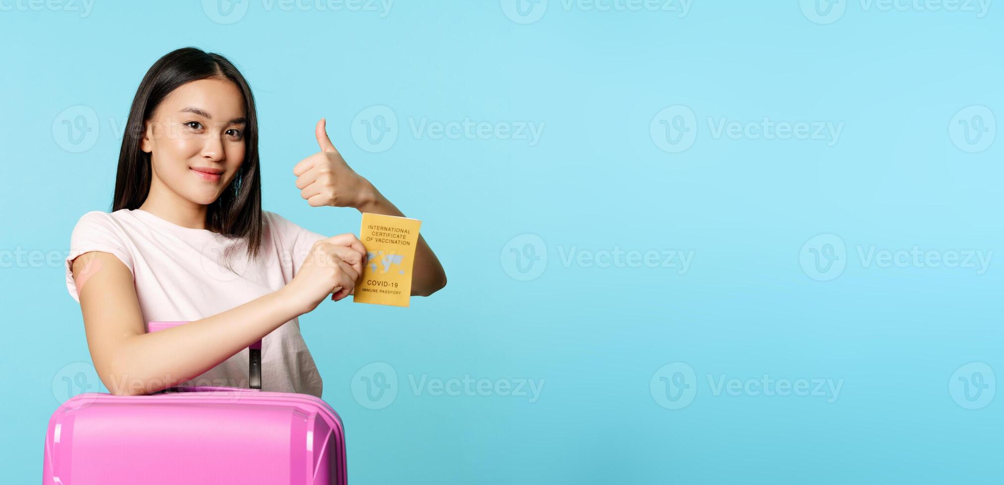 Healthcare and tourism. Smiling asian girl got vaccinated from covid-19, standing with suitcase, showing coronavirus international vaccination passport, thumbs up and arm with patch photo