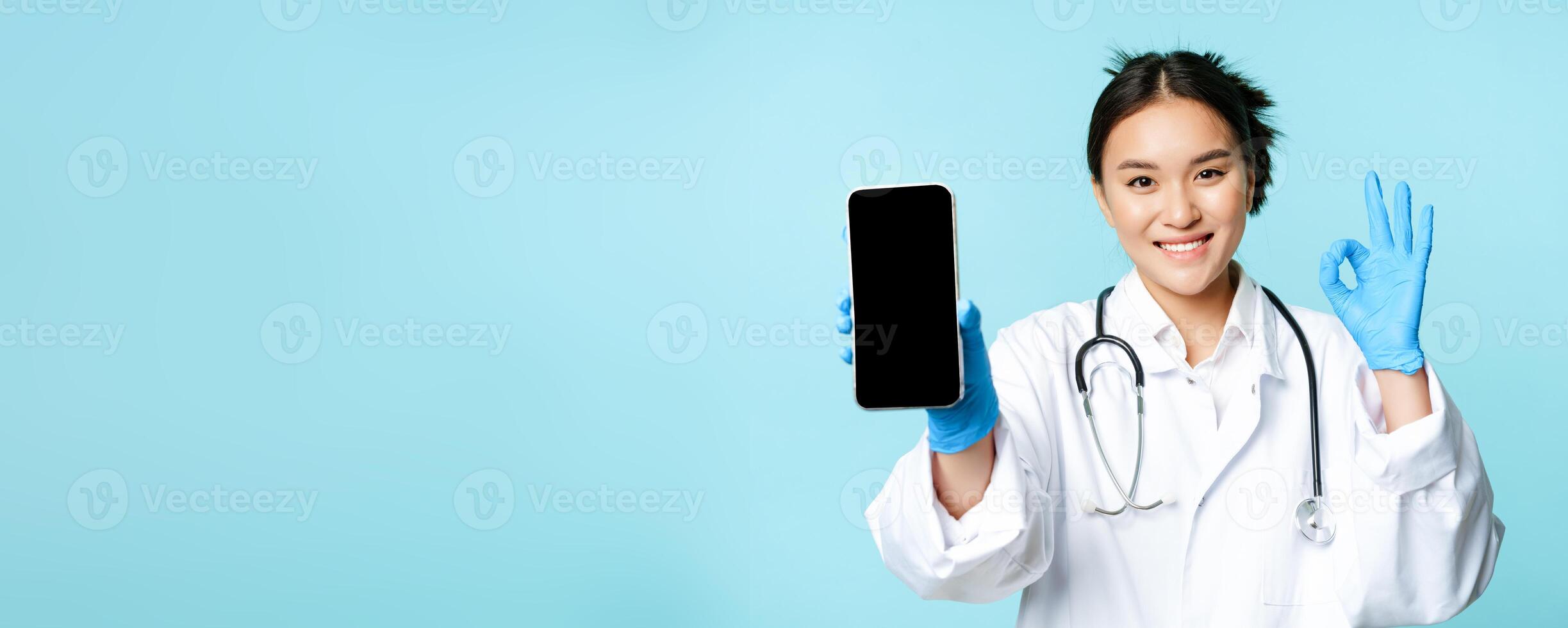 Web healthcare, online help concept. Confident asian female doctor or nurse, showing smartphone screen, app interface and okay sign in approval, recommending website photo