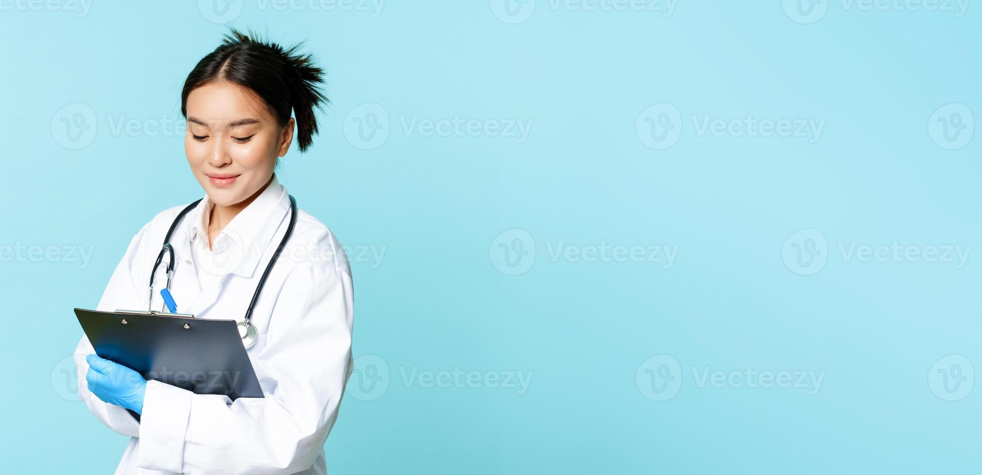 Asian female nurse or doctor writing down on clipboard with pen, smiling while examining patients, standing in uniform over blue background photo