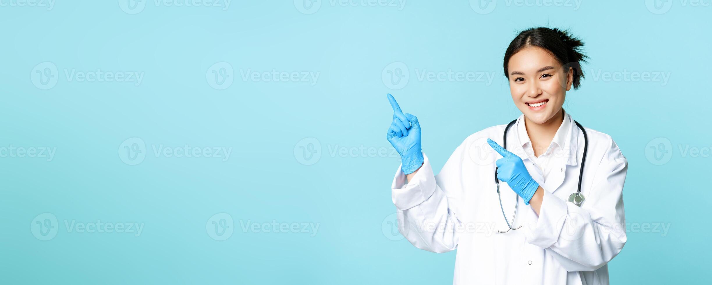 Smiling asian female doctor, therapist pointing fingers at upper left corner, showing medical advertisement, standing over blue background photo