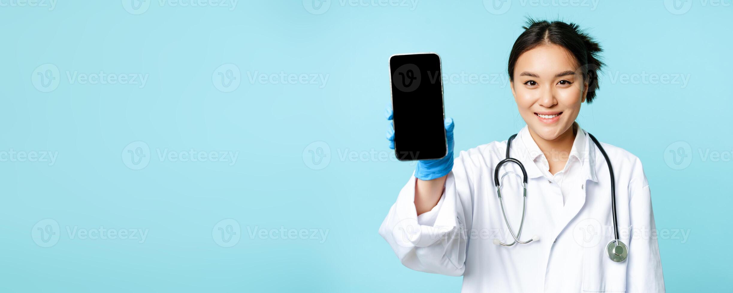 Web healthcare, online help concept. Smiling asian woman doctor, showing smartphone app interface, mobile screen, standing in uniform over blue background photo