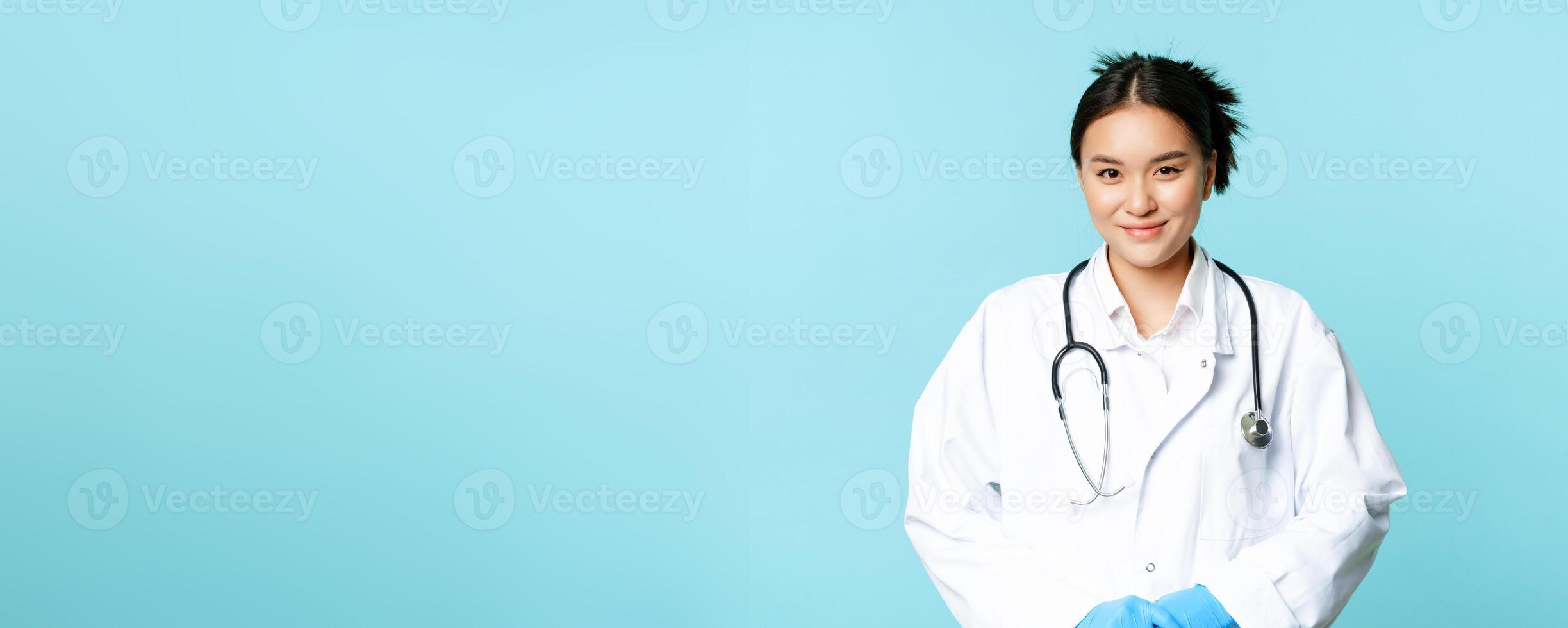 Healthcare and medical concept. Smiling asian female doctor in hospital uniform and stethoscope, looking pleasant at patient, blue background photo