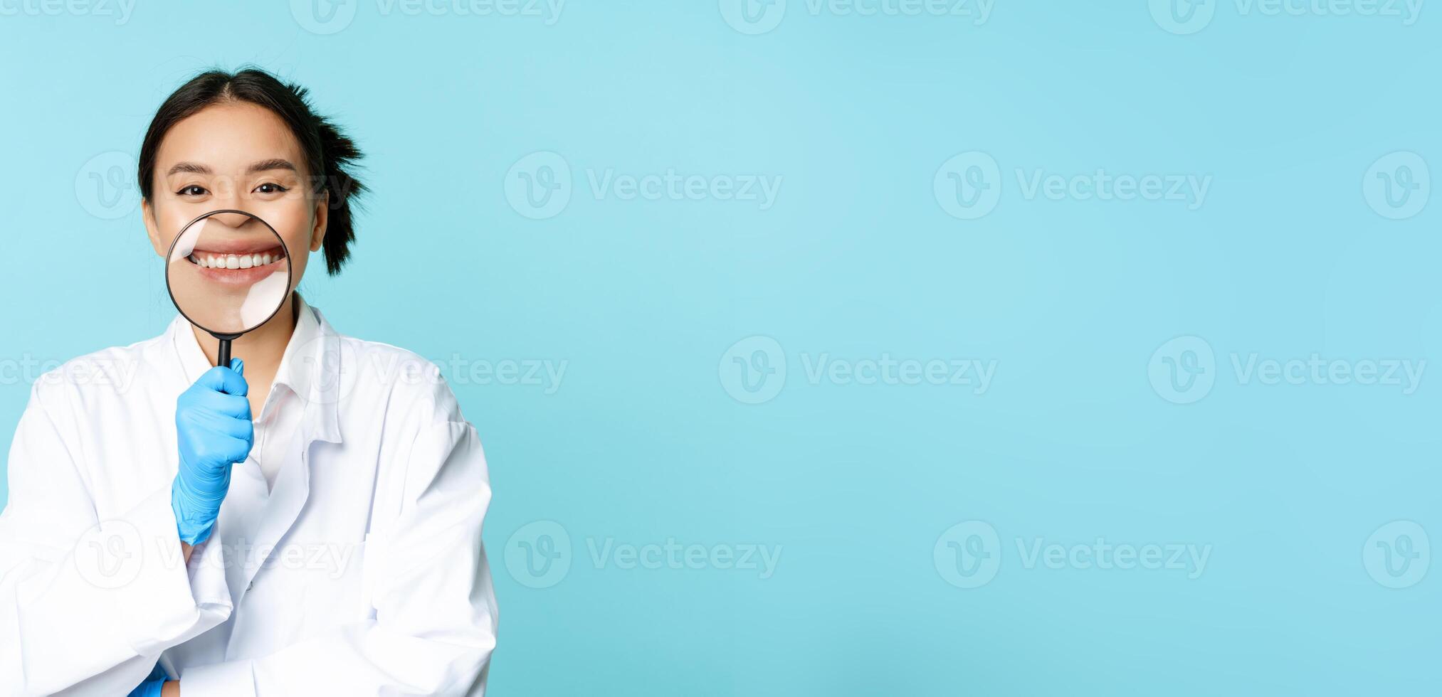 Funny asian female physician, doctor showing her smile with magnifying glass, standing in medical uniform over blue background photo