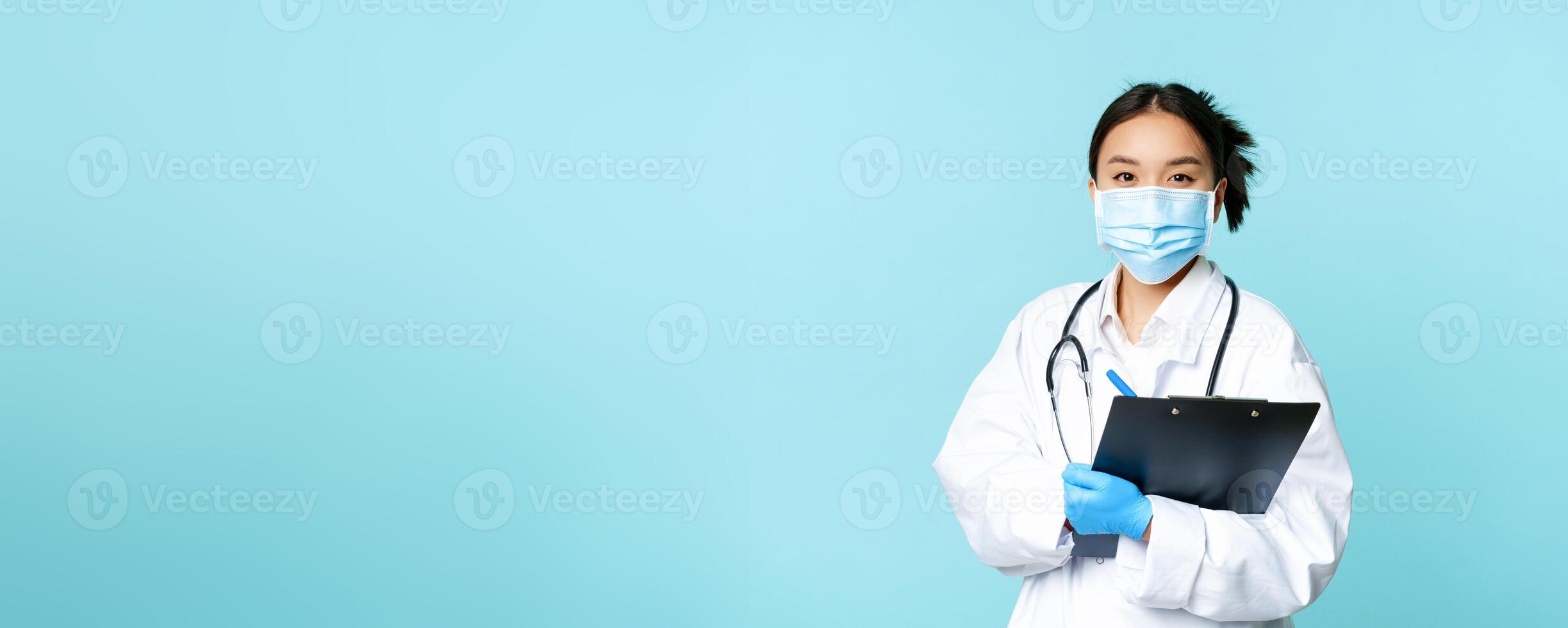 Covid-19 and quarantine concept. Smiling asian female physician, nurse in face mask holding clipboard, shift at hospital, standing over blue background photo