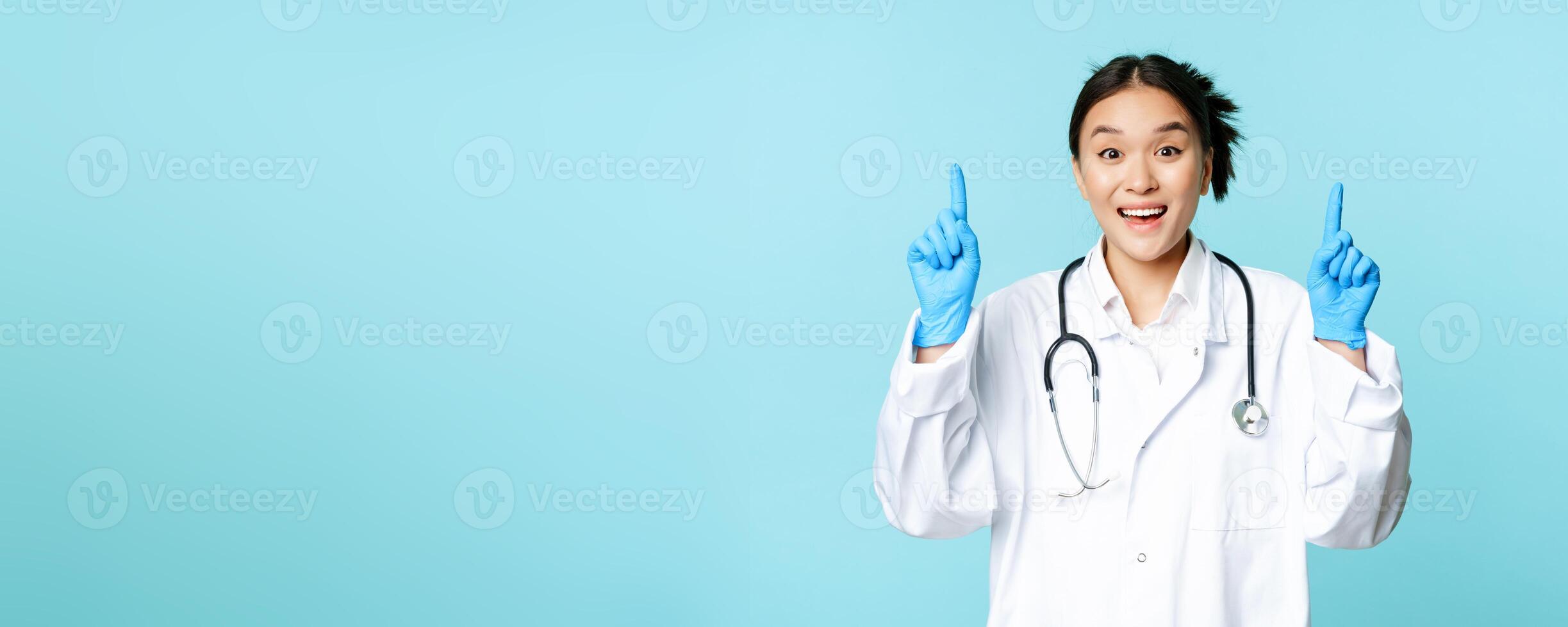 Enthusiastic asian doctor, female physician pointing fingers up and smiling, showing advertisement, demonstrating promo, blue background photo
