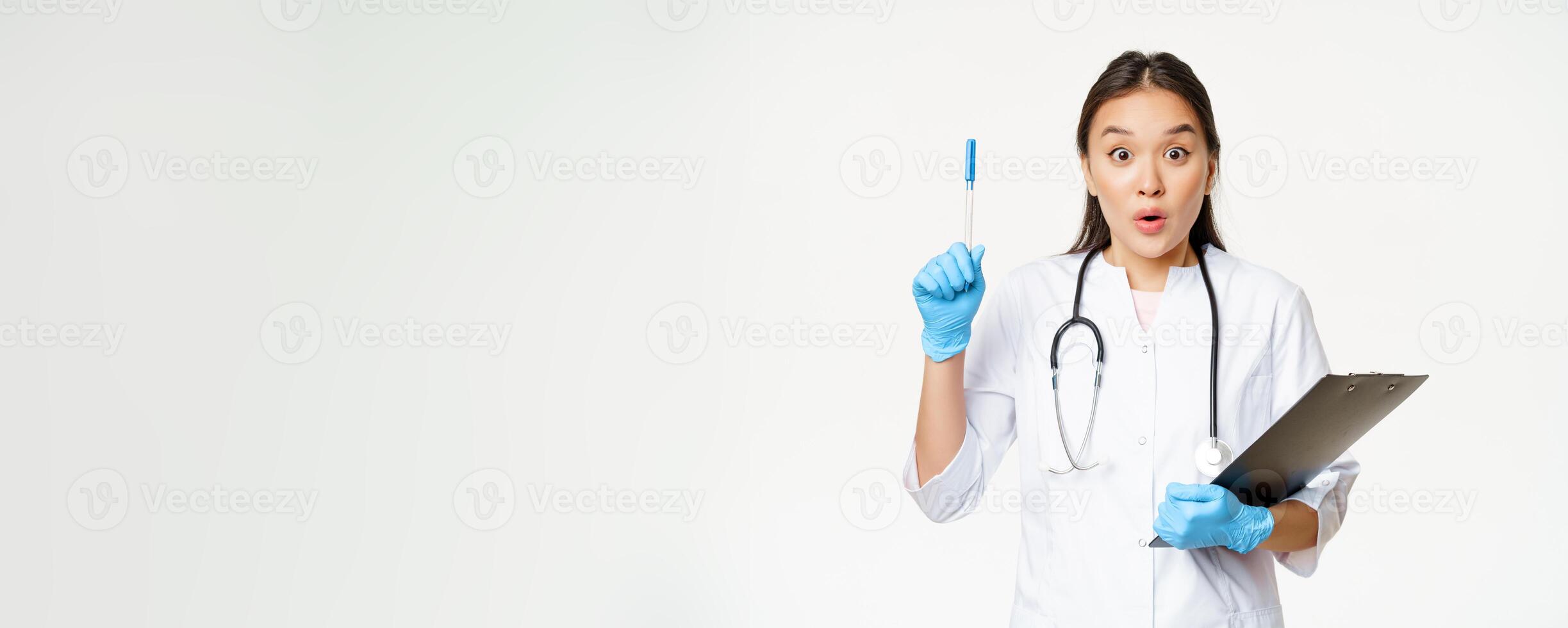 Enthusiastic nurse, asian woman doctor raising pen up, holding clipboard with patient papers, standing in uniform over white background photo