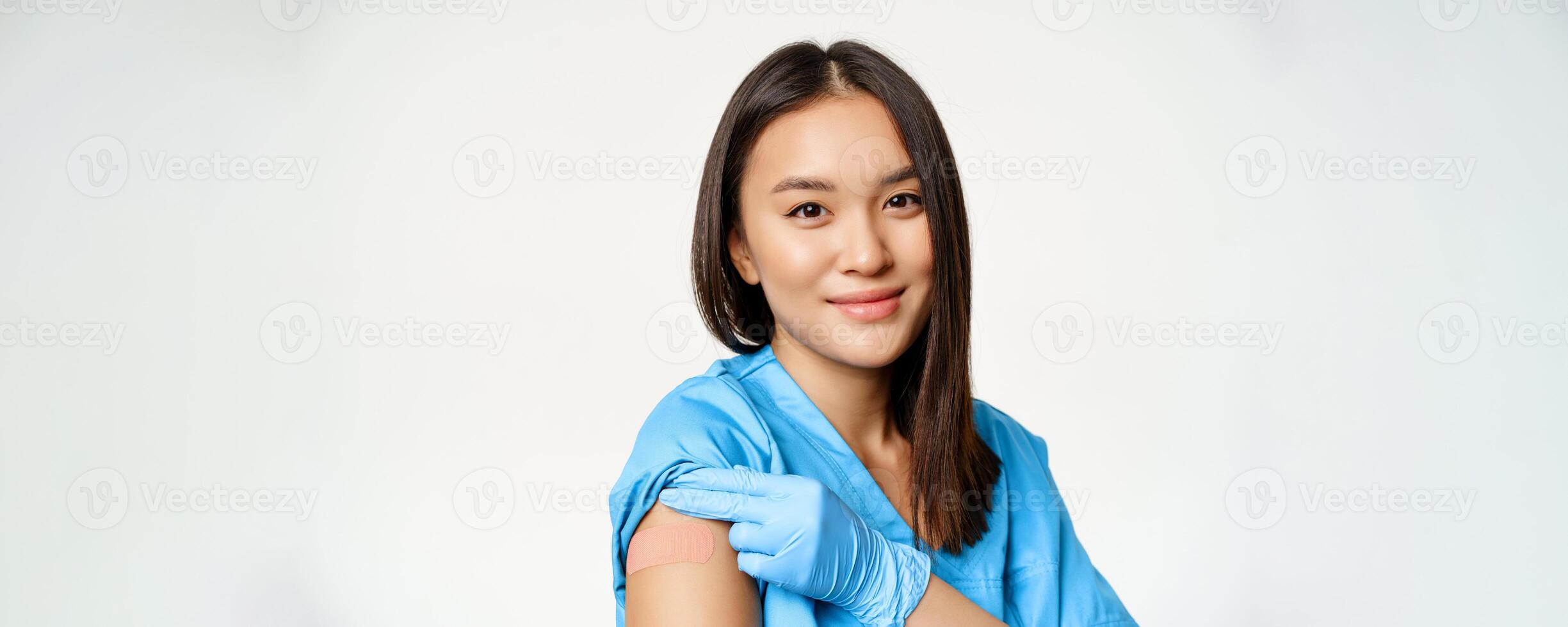 Vaccination program and covid-19 concept. Portrait of asian healthcare worker in medical robe, showing vaccinated arm and smiling, standing over white background photo