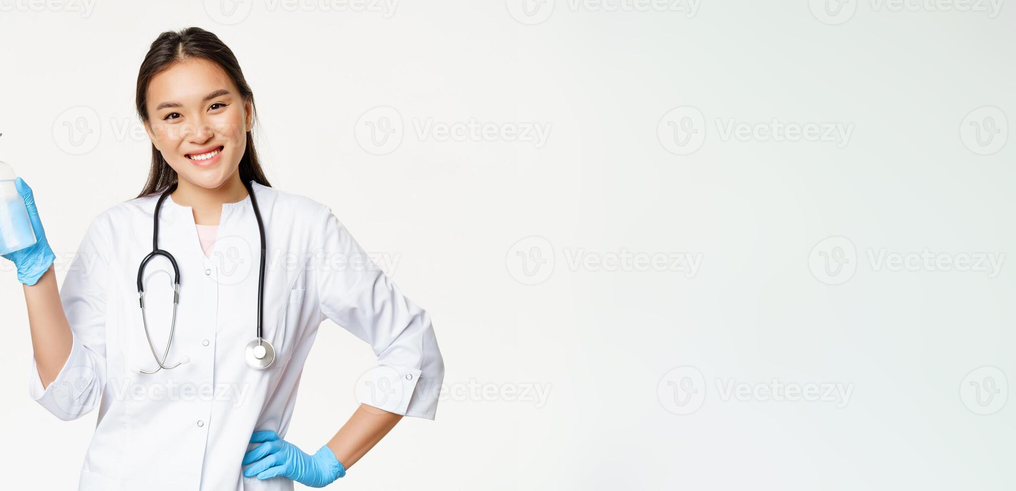 Smiling asian physician holding hand sanitizer in rubber gloves, showing bottle with antiseptic for coronavirus prevention, white background photo