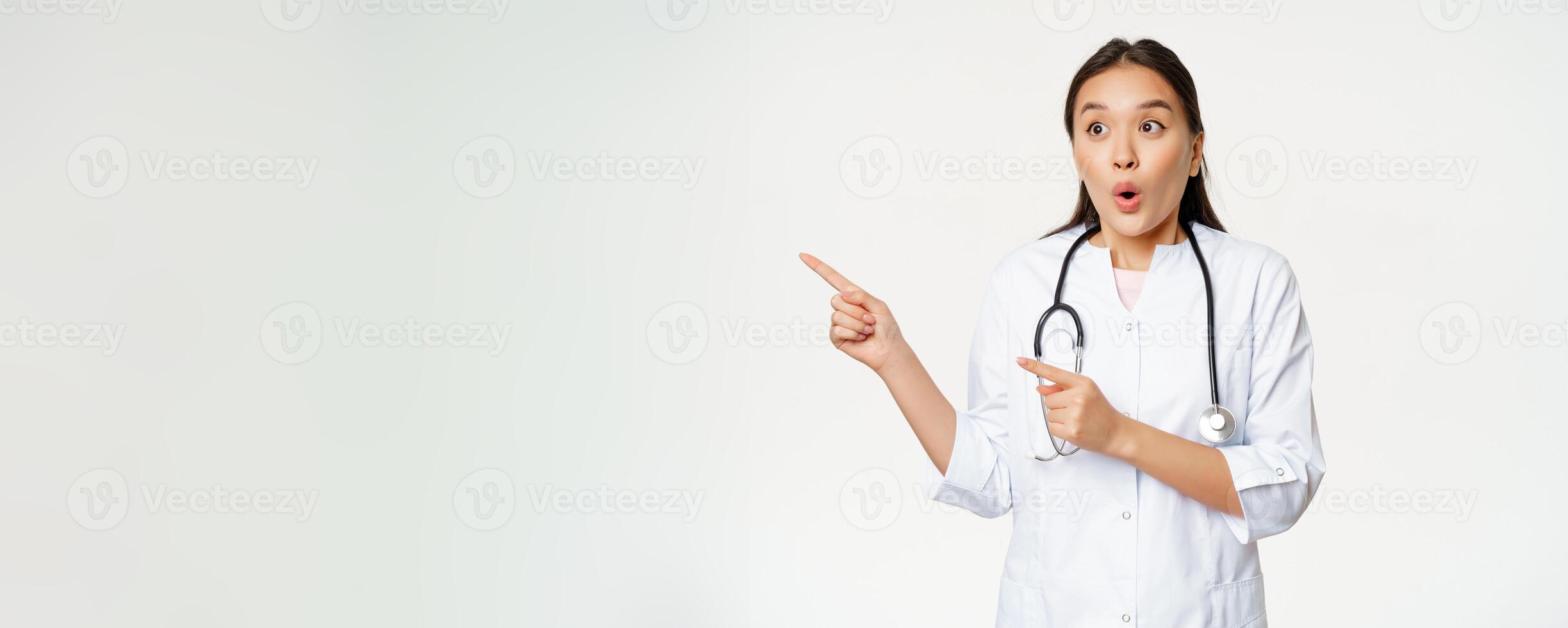 Portrait of enthusiastic female doctor, asian physician pointing and looking left with surprised, amazed face expression, standing in medical robe against white background photo