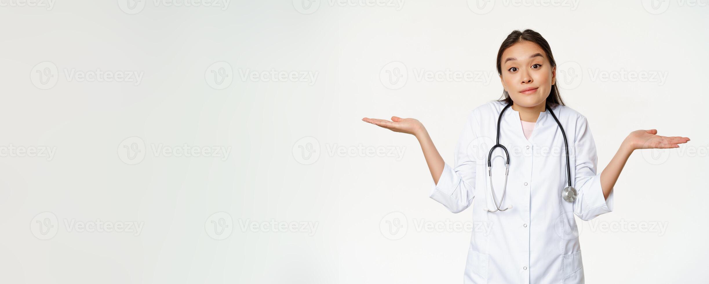 Dont know. Clueless female asian doctor shrugging, spread hands sideways and looking puzzled, cant understand, standing over white background photo