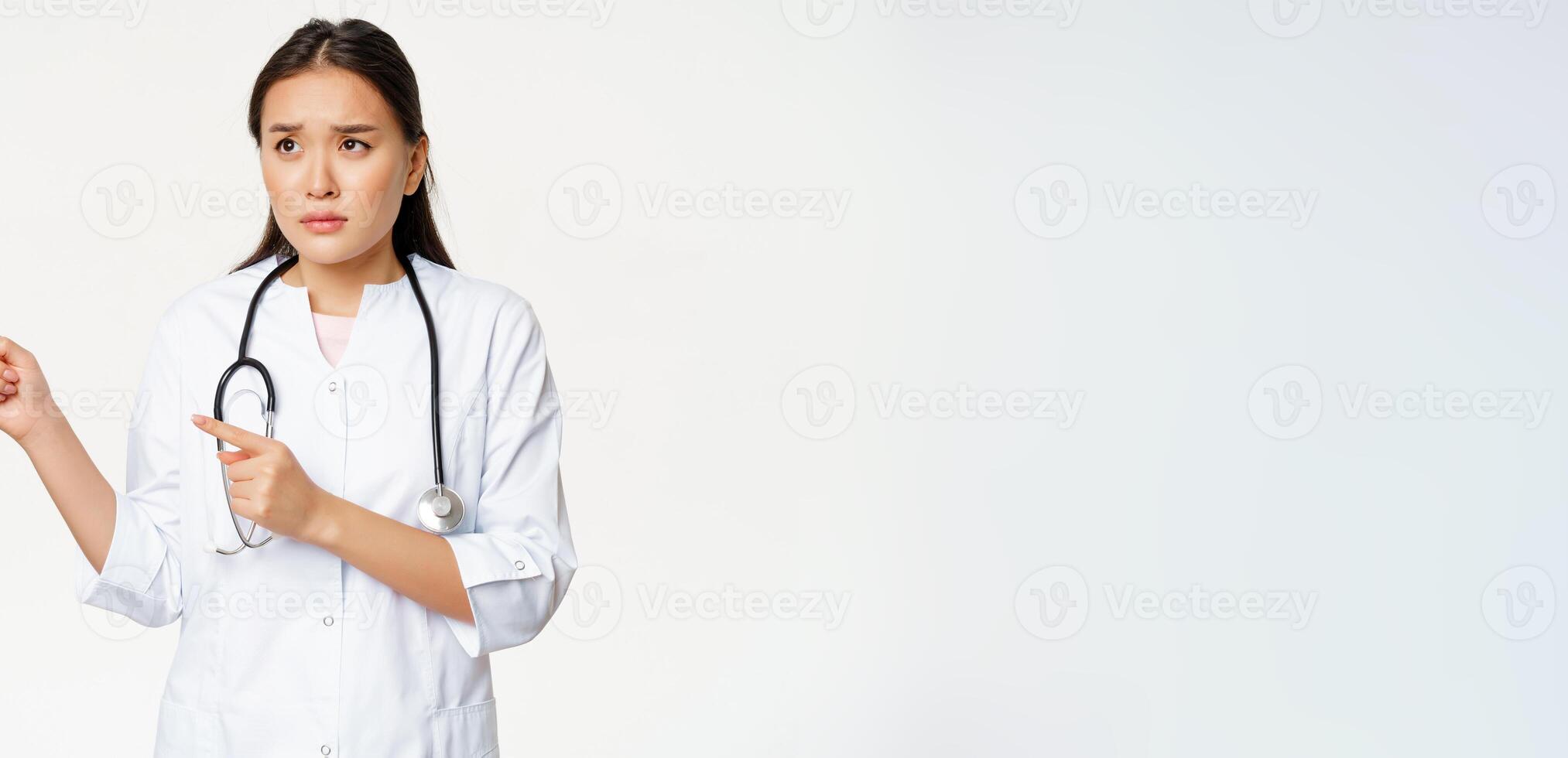 Skeptical female doctor, concerned nurse pointing and looking left with worried face expression, standing in white medical robe against studio background photo