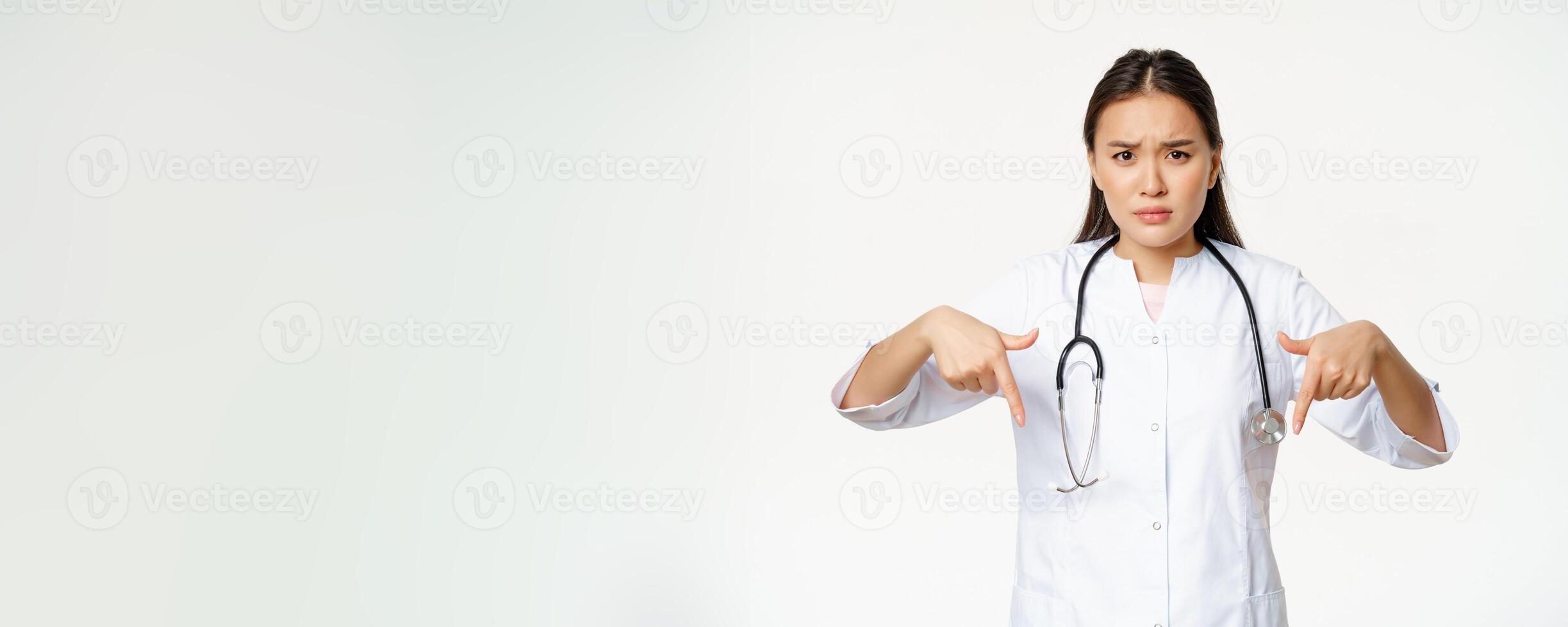 Disappointed asian female doctor pointing fingers down, frowning and looking concerned, showing bad product, standing over white background photo