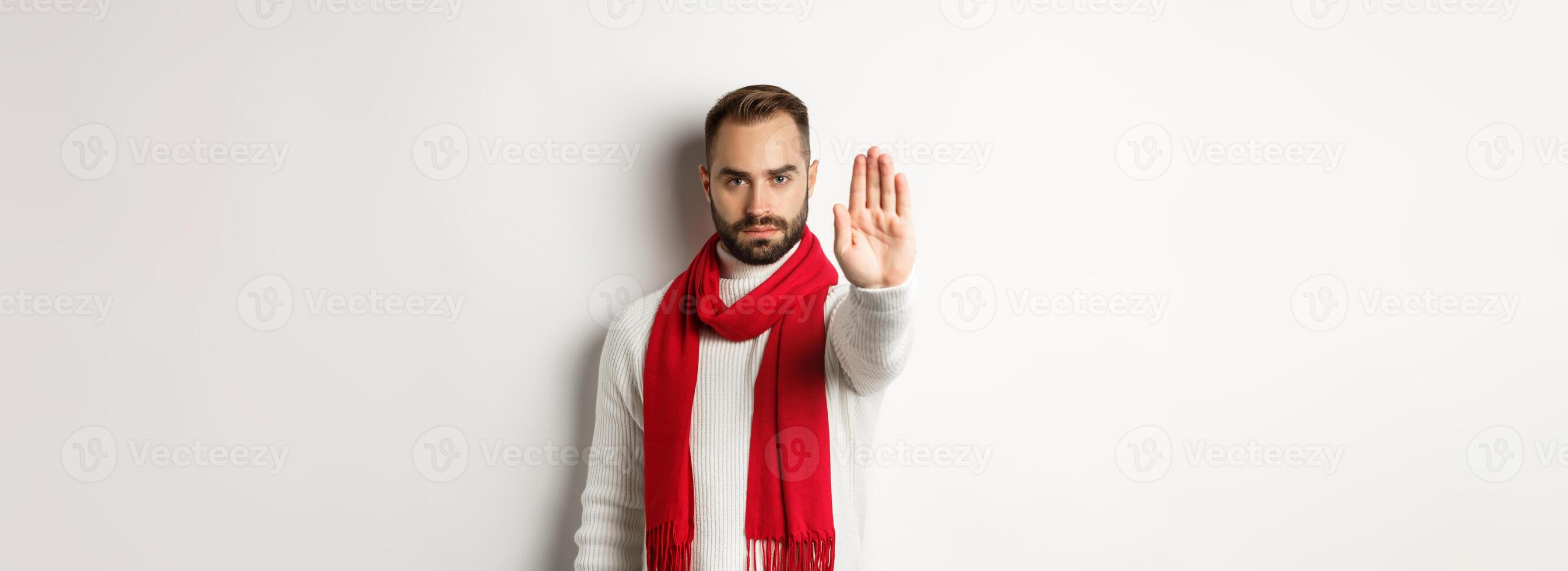 Serious bearded guy saying no, showing stop sign, rejection sign, prohibit action, standing in winter sweater and red scarf against white background photo