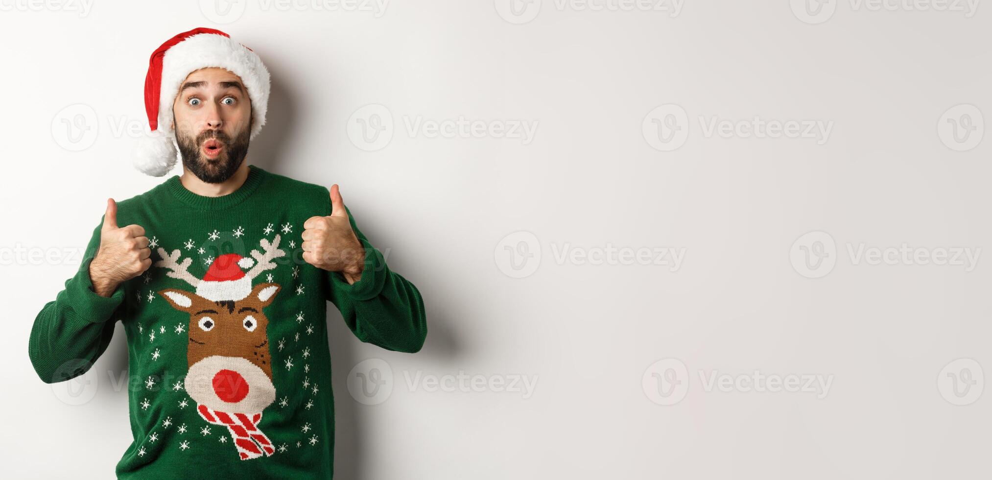 Christmas holidays, celebration and party concept. Amazed young man in Santa hat and sweater, showing thumb up in approval, like something, white background photo