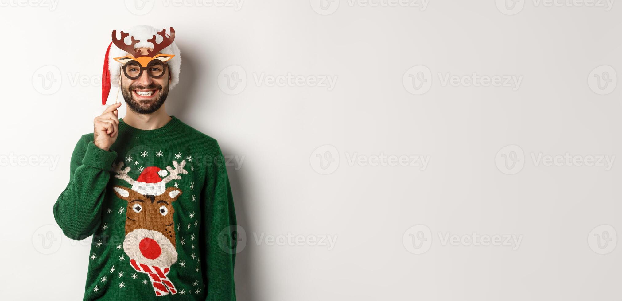 Christmas and holidays concept. Smiling beared man in Santa hat looking happy, holding party mask for New Year, celebrating xmas, standing over white background photo