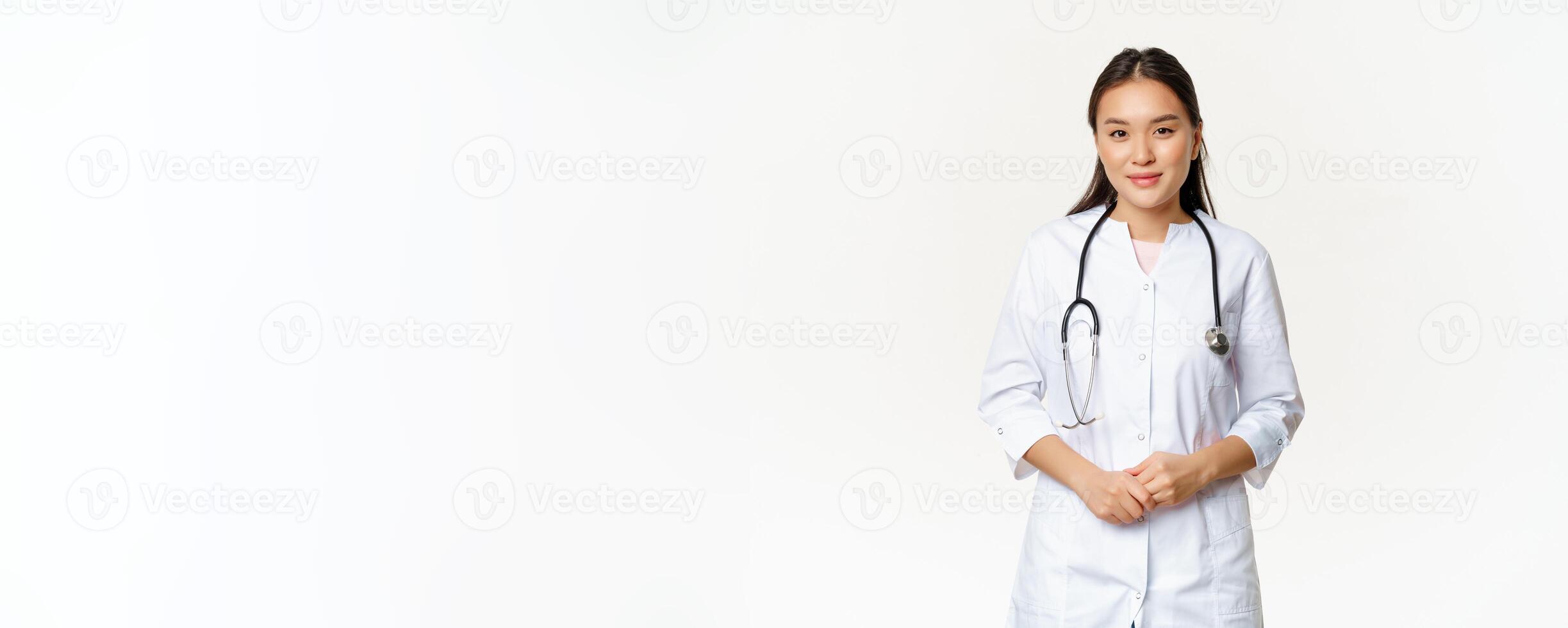 Smiling asian medical worker with stethoscope, wearing doctor uniform, looking helpful at patient, standing over white background photo