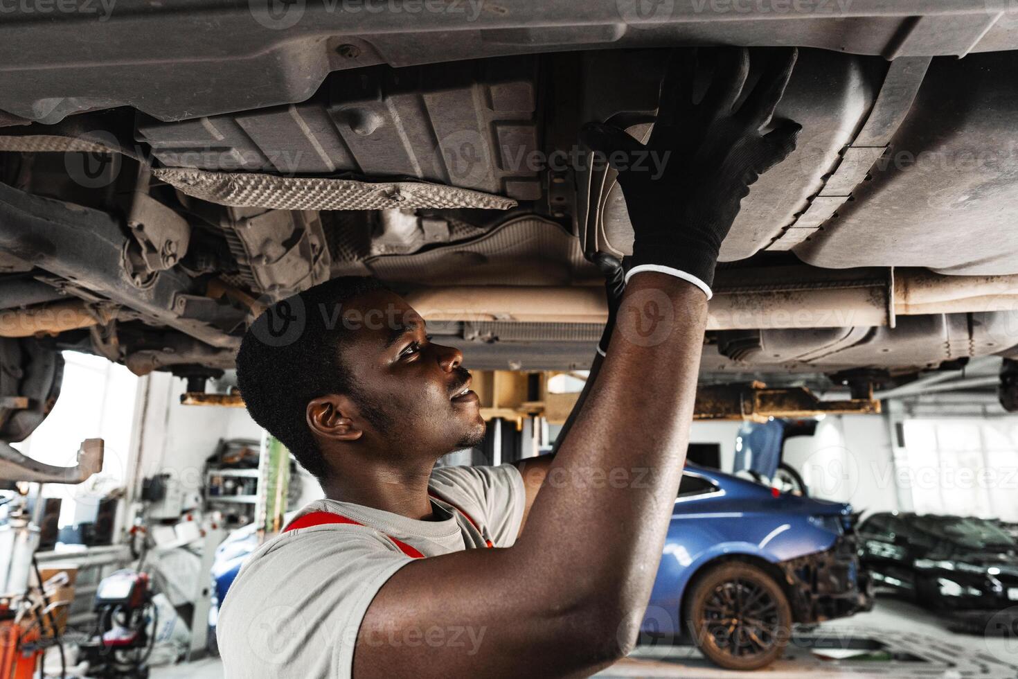 Young African mechanic in uniform working under the car in car service center photo