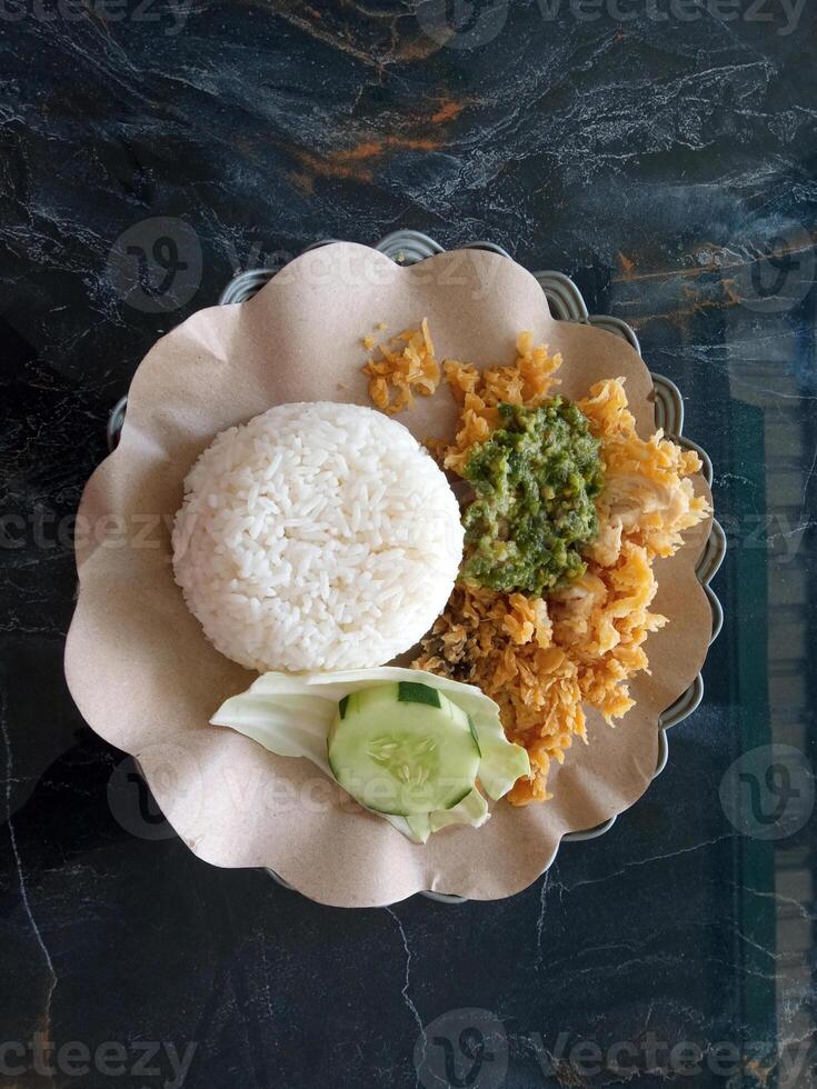 Top view of a plate of rice and ayam geprek, literally translated smashed chicken, with a slice of cucumber, cabbage and green chili sauce. photo