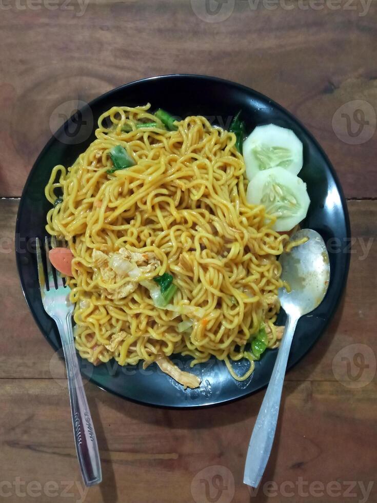 Flat lay view of delicious fried noodles with some slices of fresh cucumber on a black ceramic plate. photo