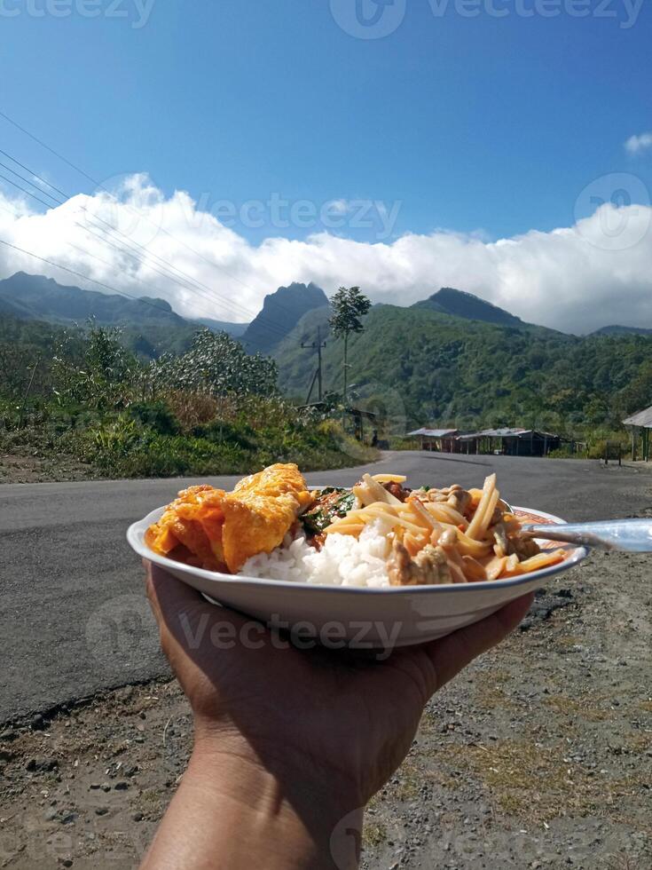 Outdoor breakfast with mountain view. Someone is holding a plate of rice with beautiful scenery in the background. photo