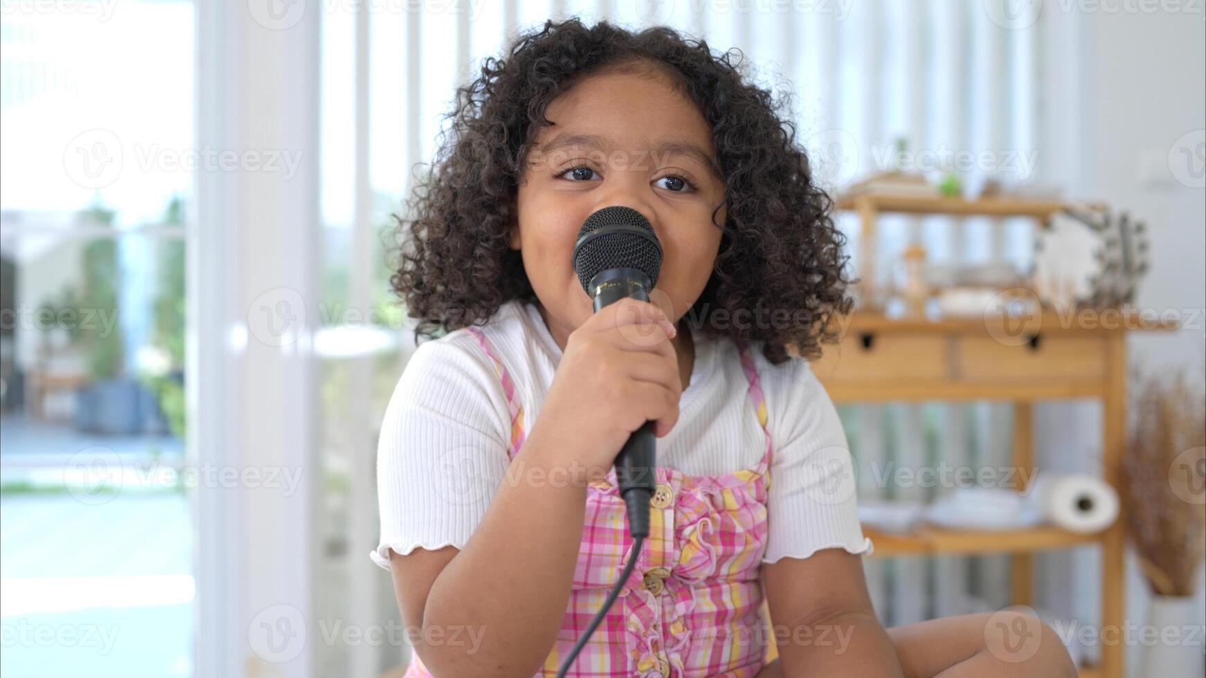 A tiny girl sits on a toy table, clutching a microphone, and sings cheerfully in the living room photo