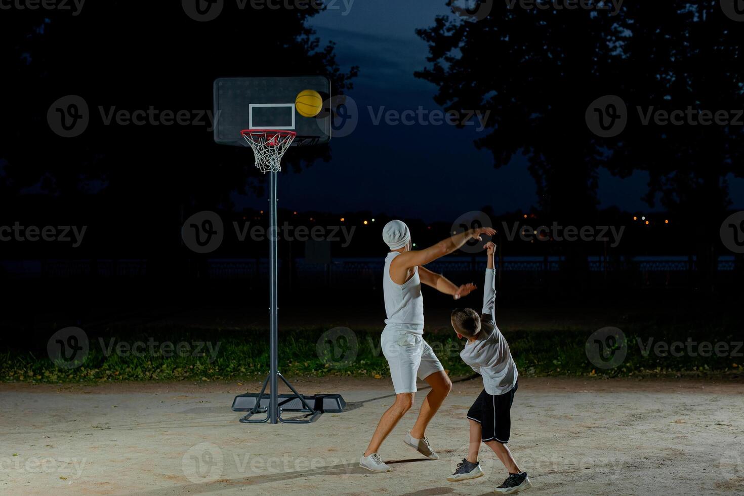 A father and son enjoy a night basketball game at a local park under bright lights focusing on teamwork and fun photo
