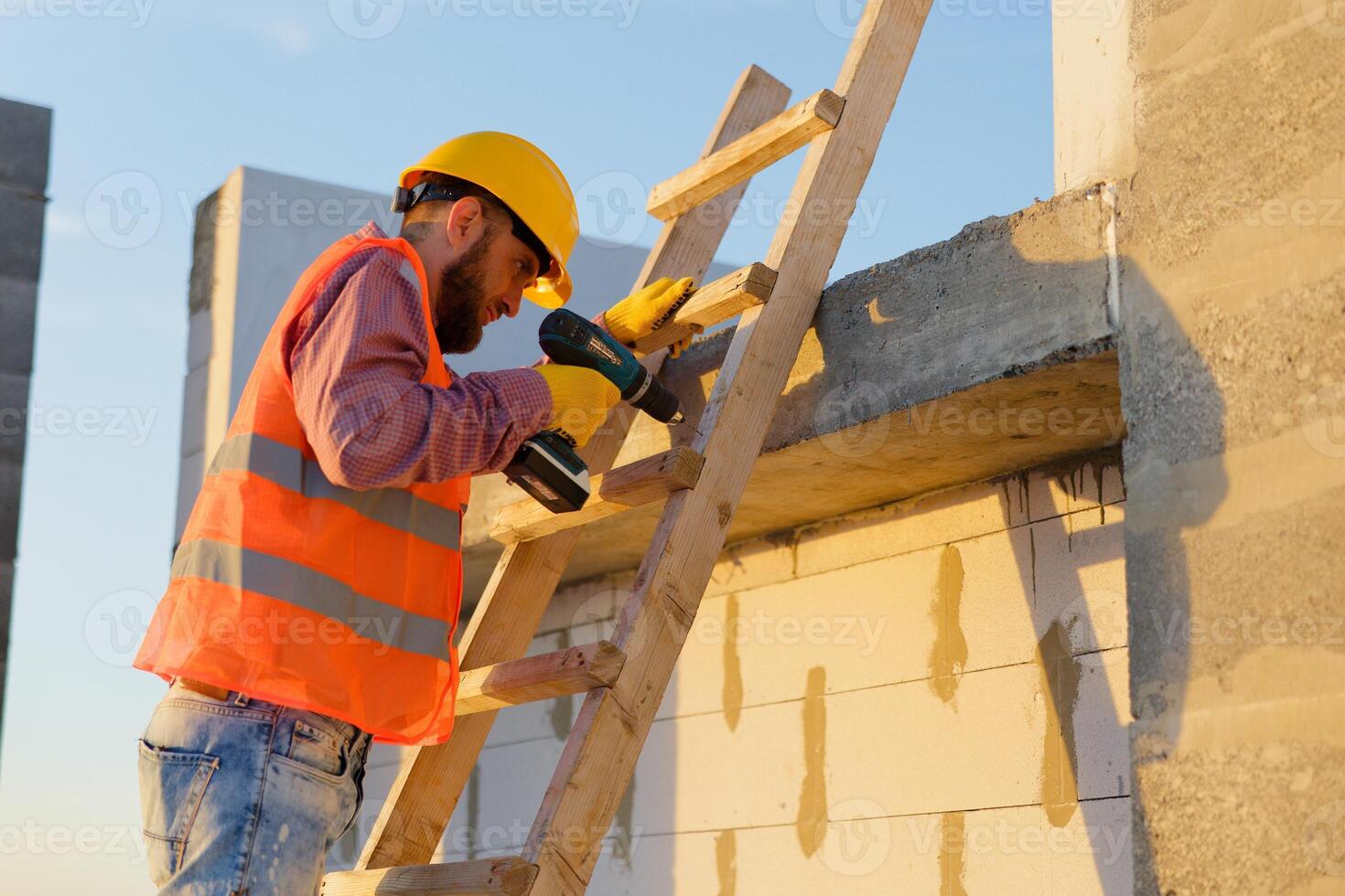 Construction worker using cordless drill on wooden ladder at building site photo
