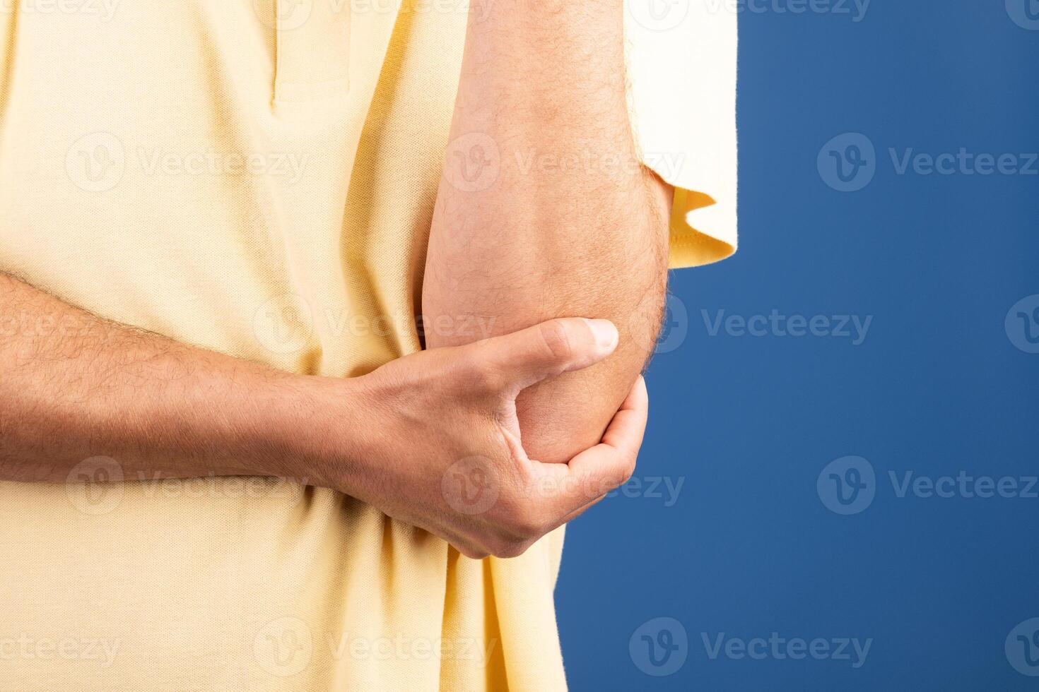 A mature Caucasian man in his 50s is sitting alone, looking uncomfortable as he holds his elbow. The background is a calming blue, suggesting a moment of solitude and reflection. photo
