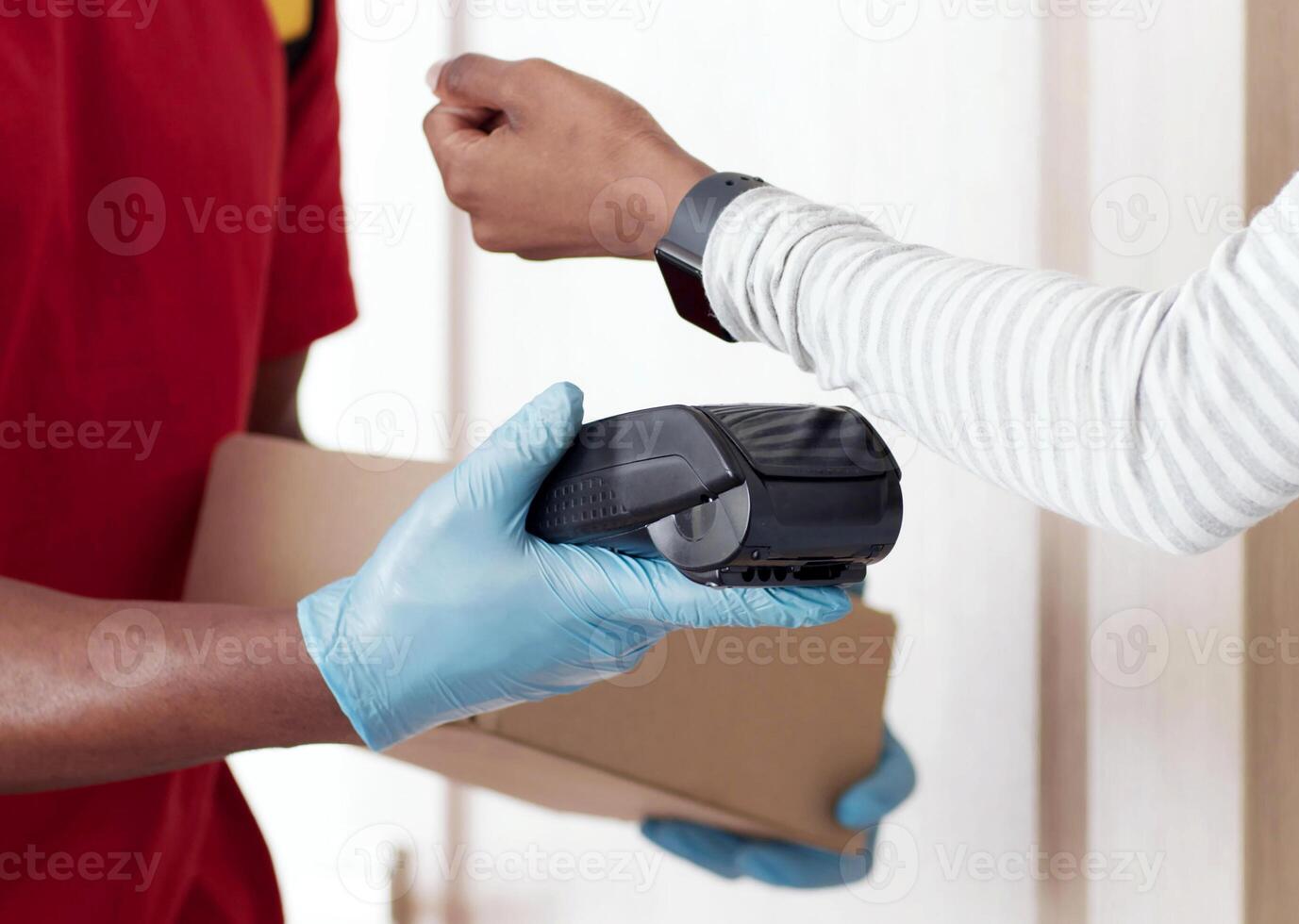Modern payment technologies and self isolation. Close up shot of young black woman paying for order with smartwatch, taking box from courier in protective gloves, covid-19 preventive measures concept photo