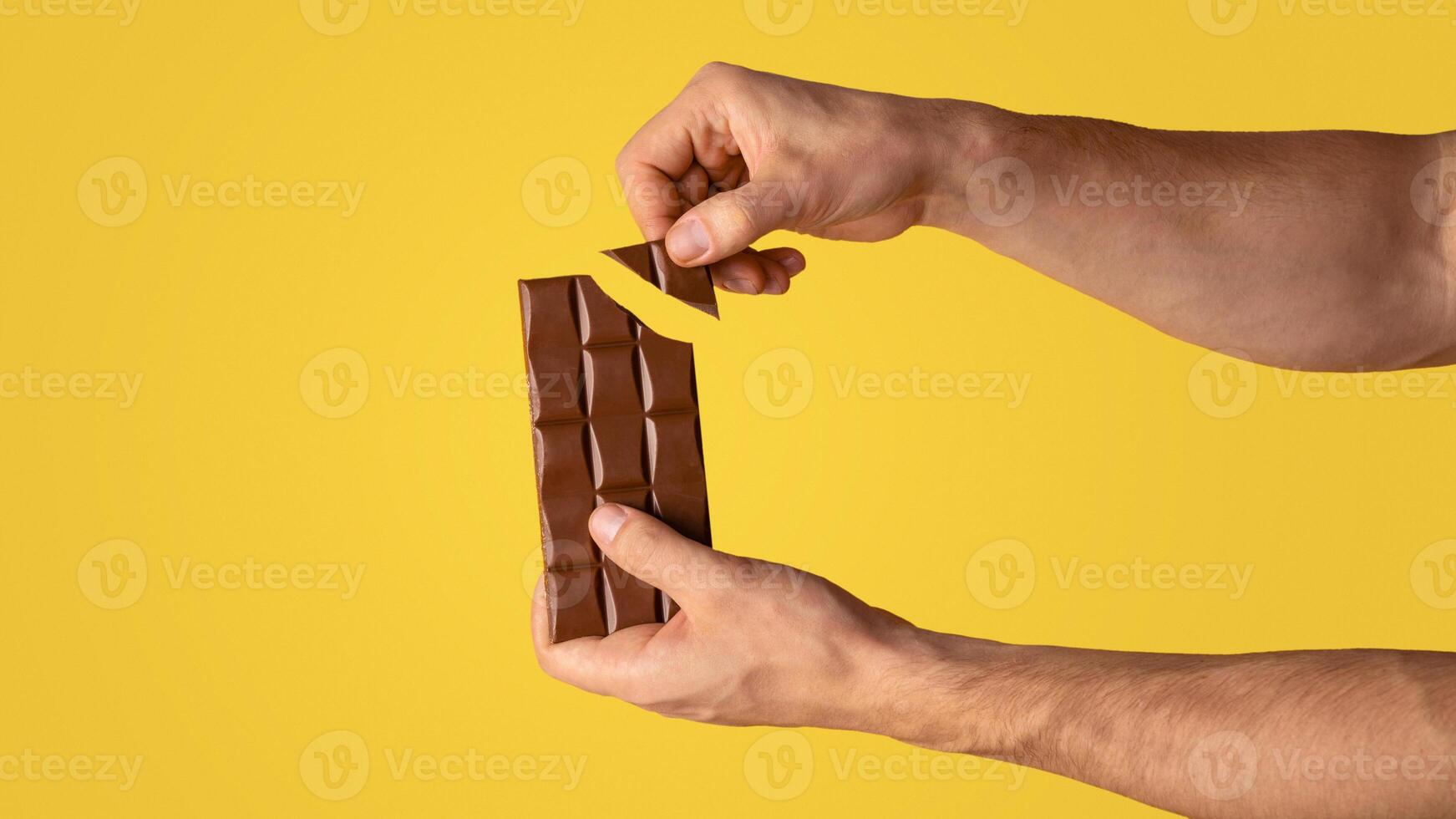 Closeup view of man breaking piece of chocolate from bar, orange background. Copy space. Panorama photo